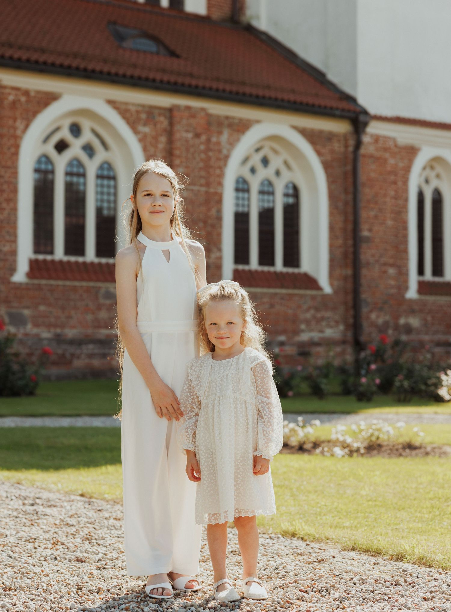Children in white dresses standing together on gravel path by brick church building.