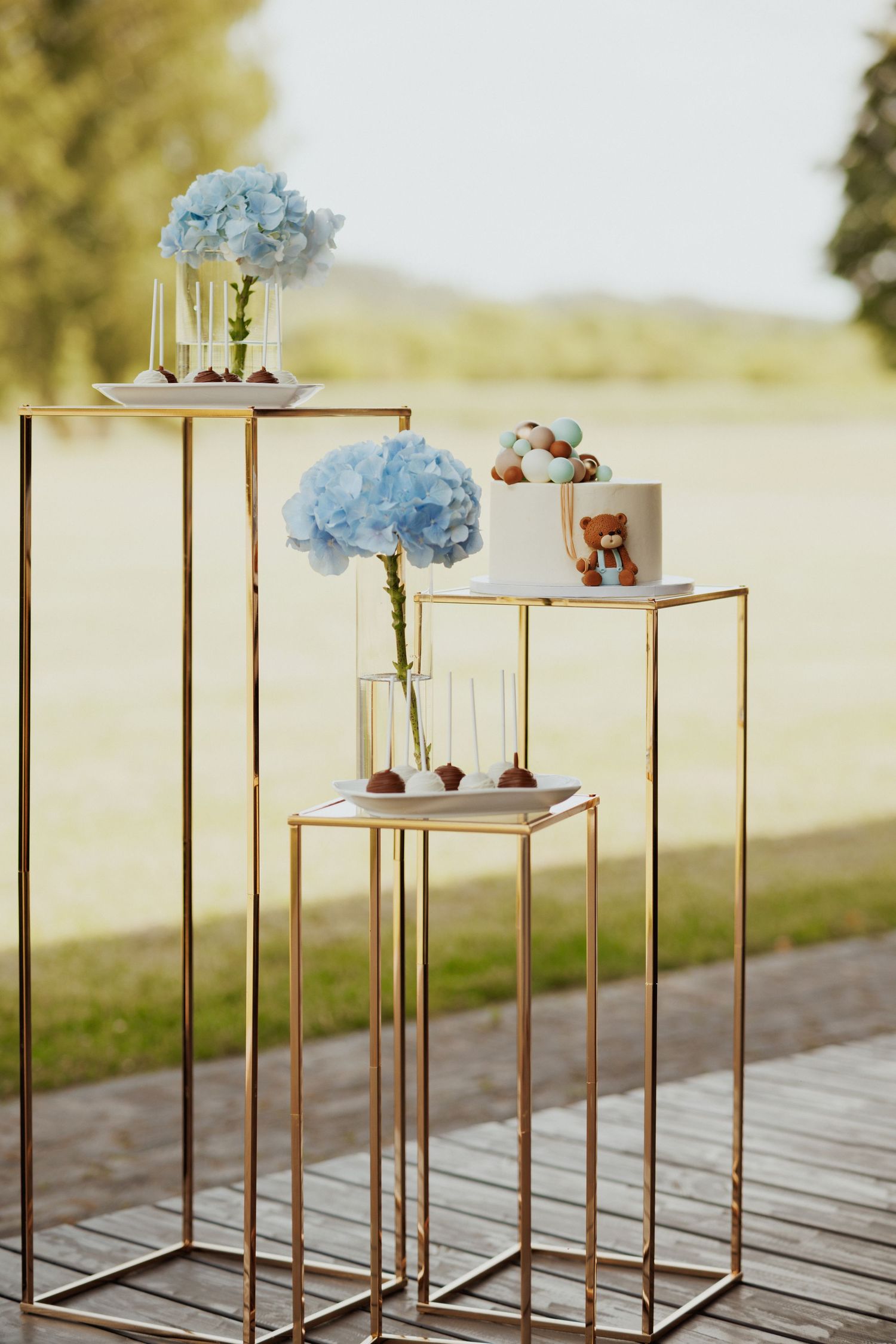 Modern gold geometric pedestals displaying desserts and blue hydrangea arrangements on an outdoor deck.