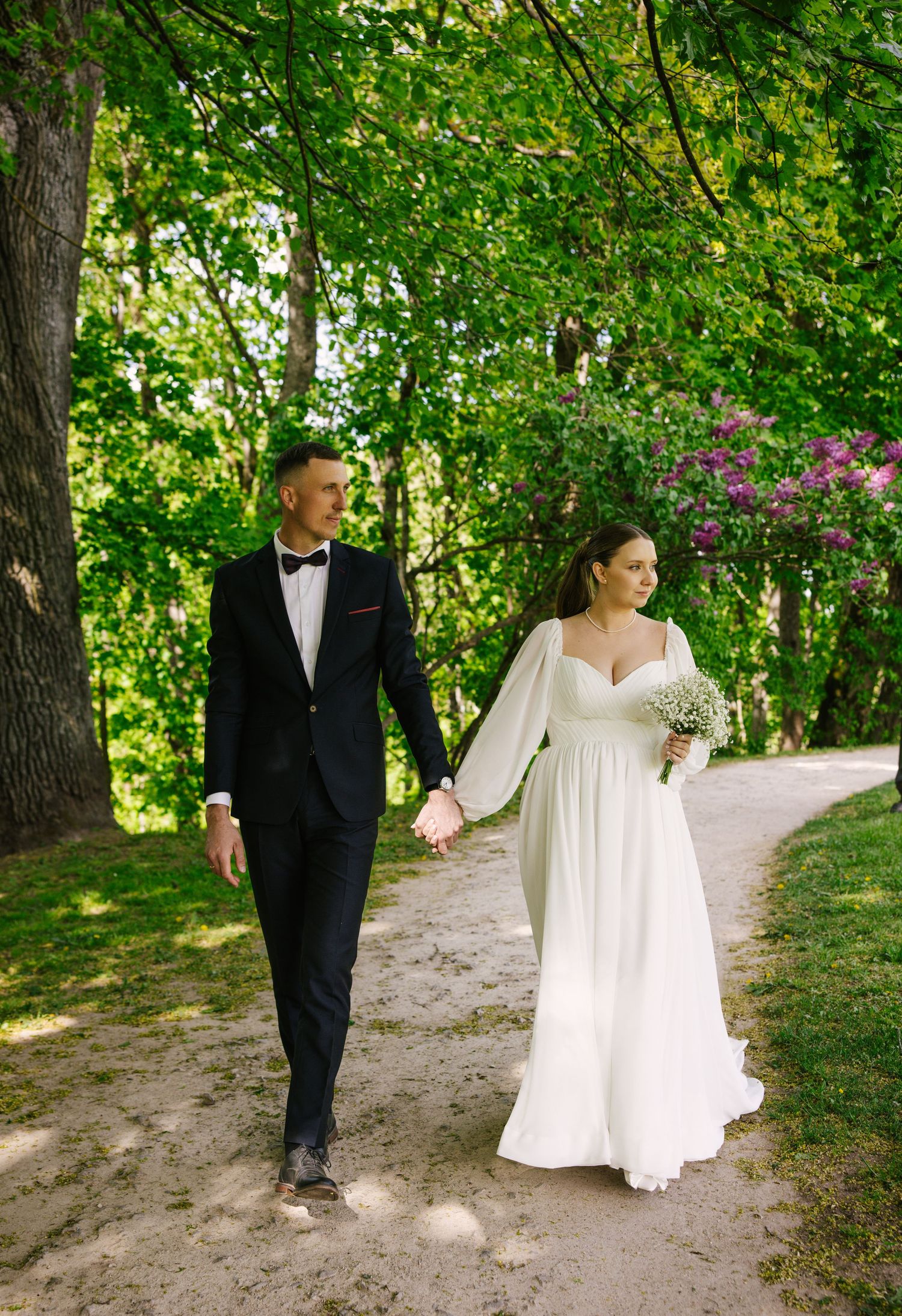 A wedding couple walks hand in hand along a garden path surrounded by lush green trees and pink flowers.