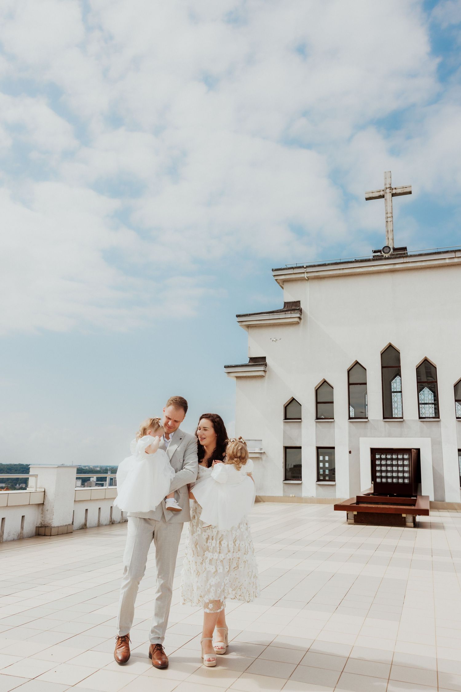 A couple shares a romantic moment on a white church rooftop terrace with a cross and blue sky in the background.