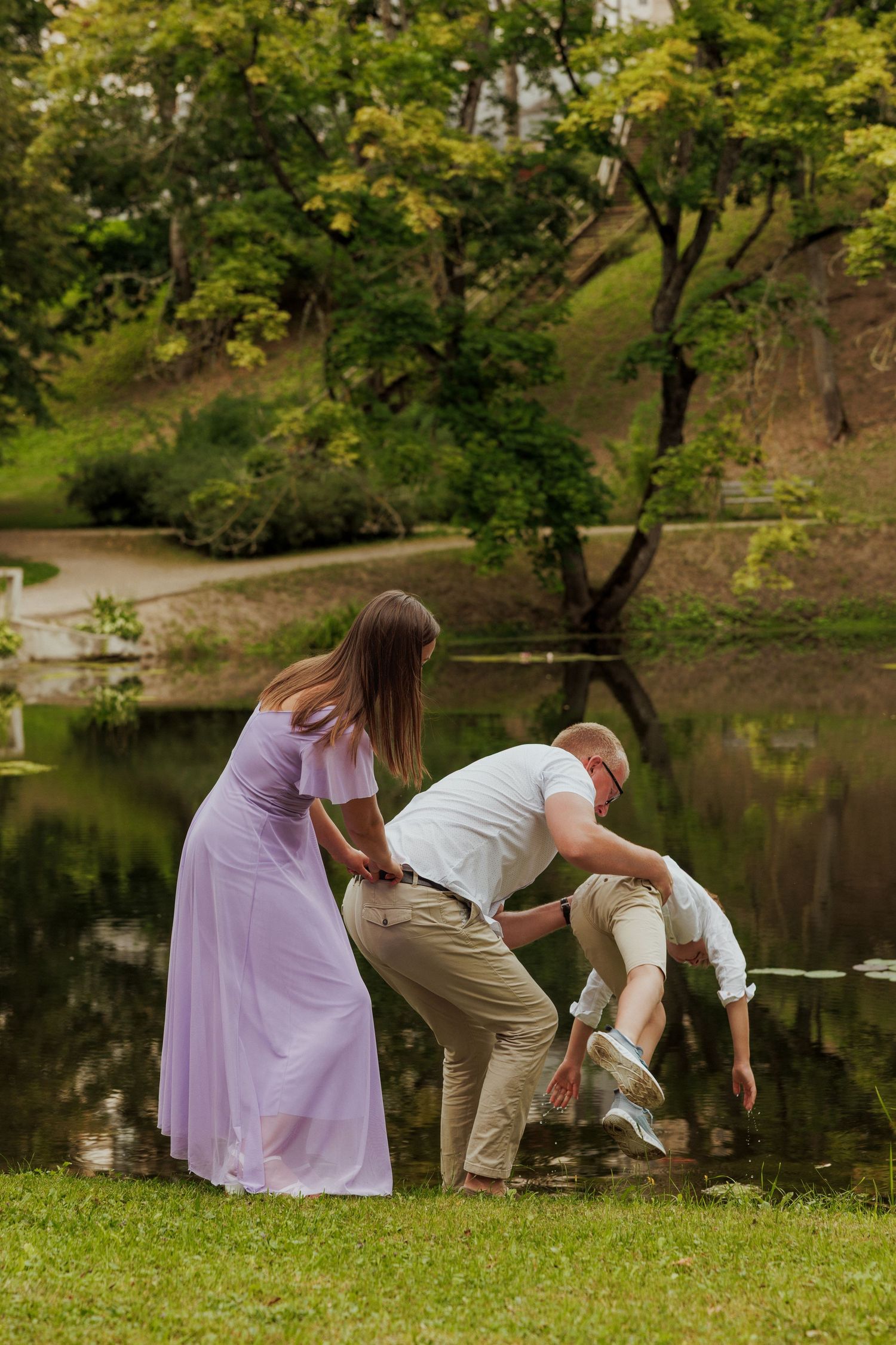 A couple in casual attire plays with a small child by a serene pond surrounded by lush green trees.