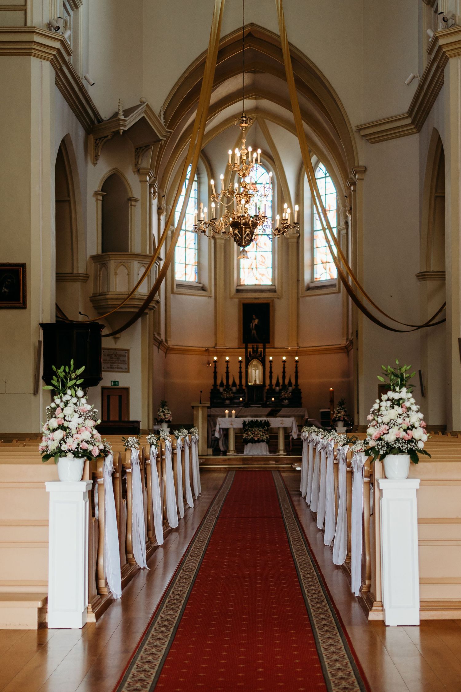 Elegant church aisle decorated with white floral arrangements and red carpet leading to illuminated altar.