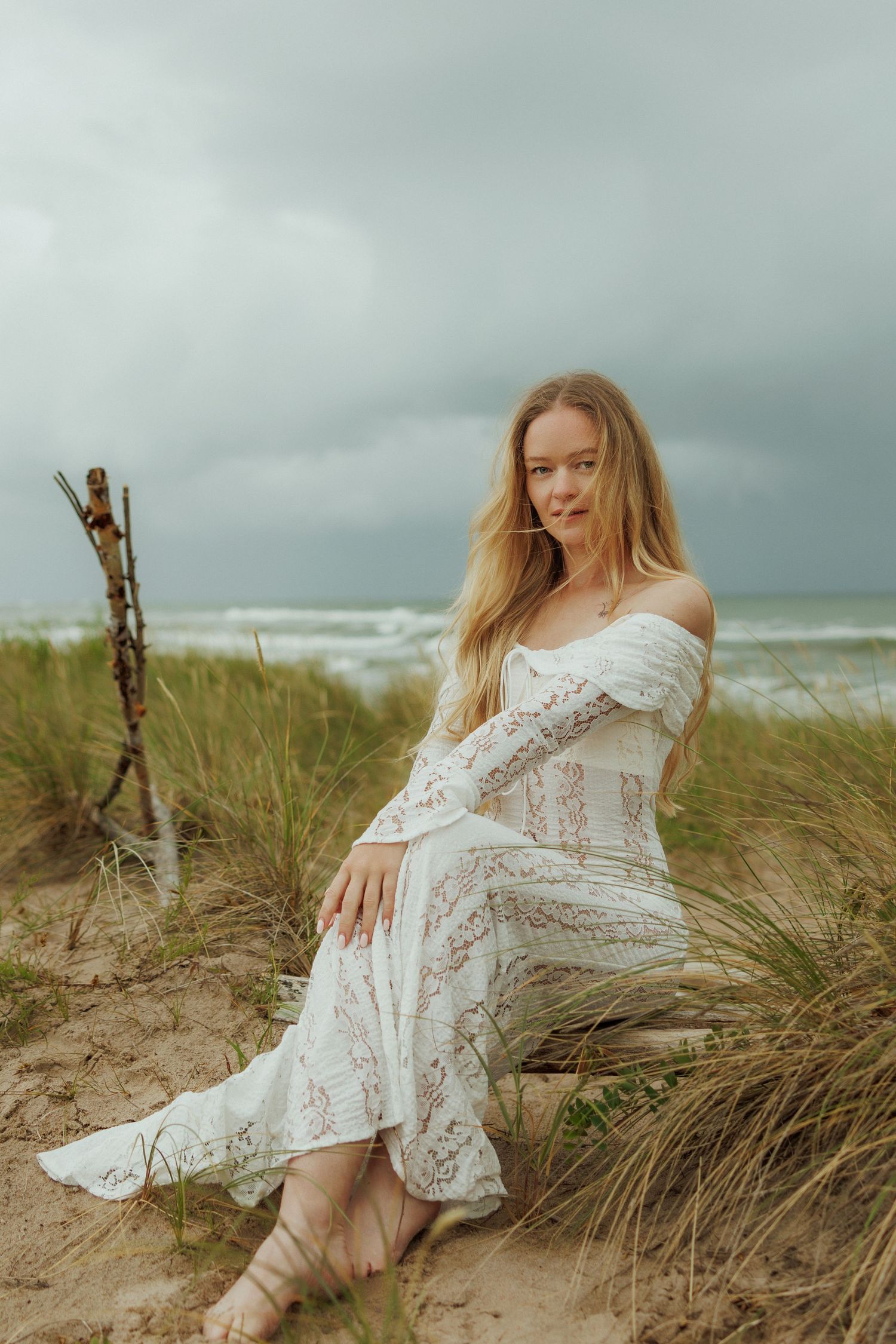 Woman in white lace dress sitting on driftwood among beach grass with stormy ocean backdrop.