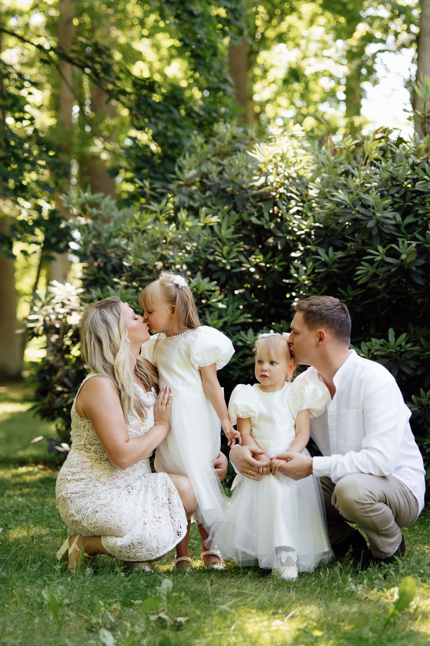 A loving family with two young daughters in white dresses share kisses in a sunlit garden setting.
