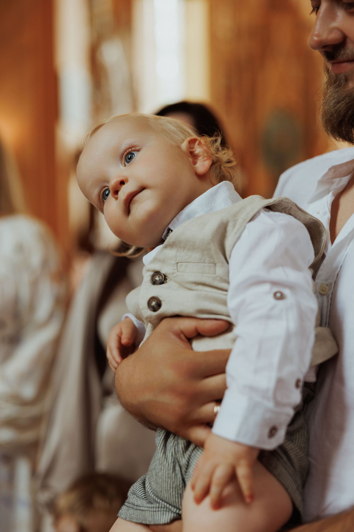 Baby in white shirt and gray overalls looks up with wonder during a formal occasion.