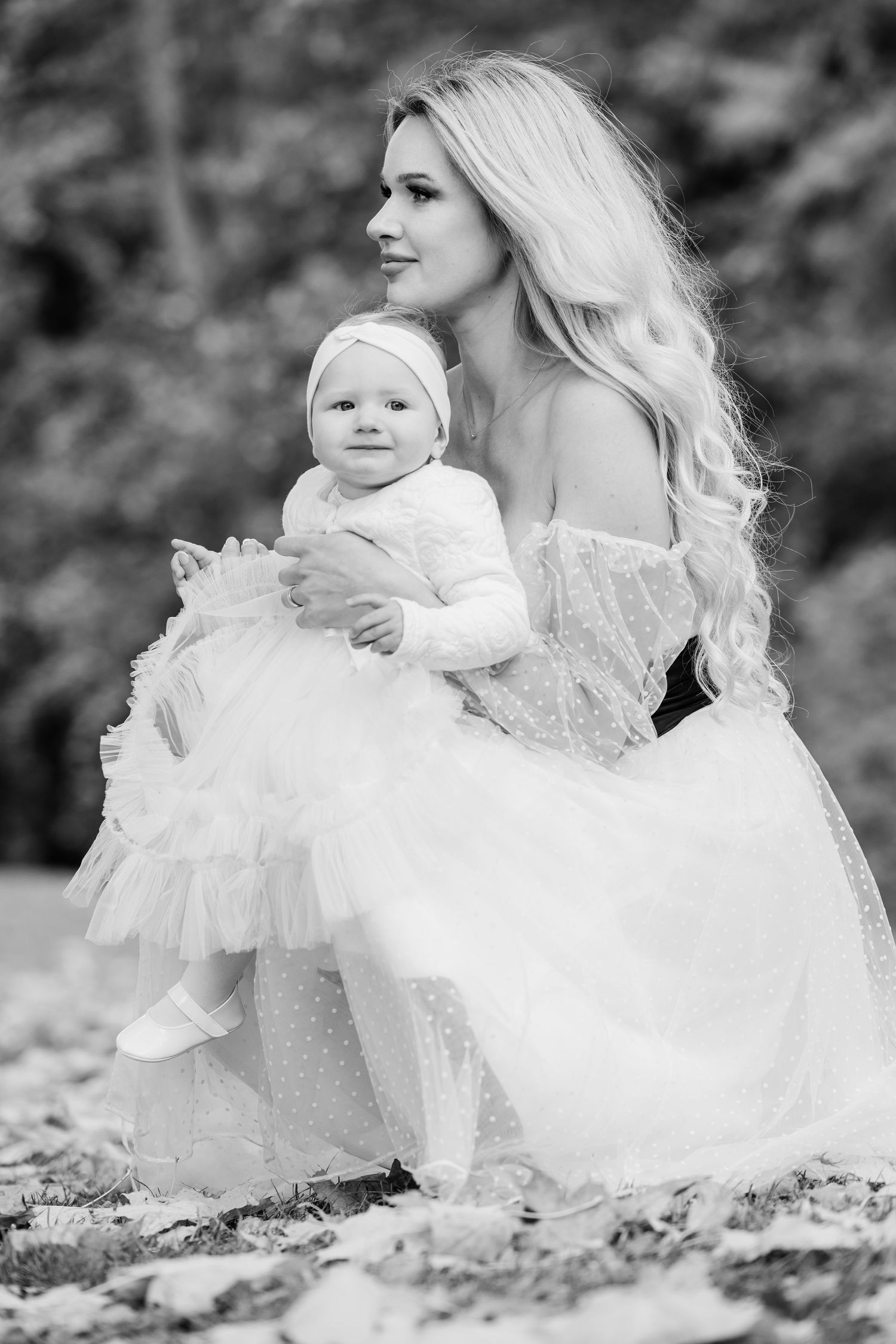 Black and white portrait of mother and baby in white dresses sitting among fallen autumn leaves.