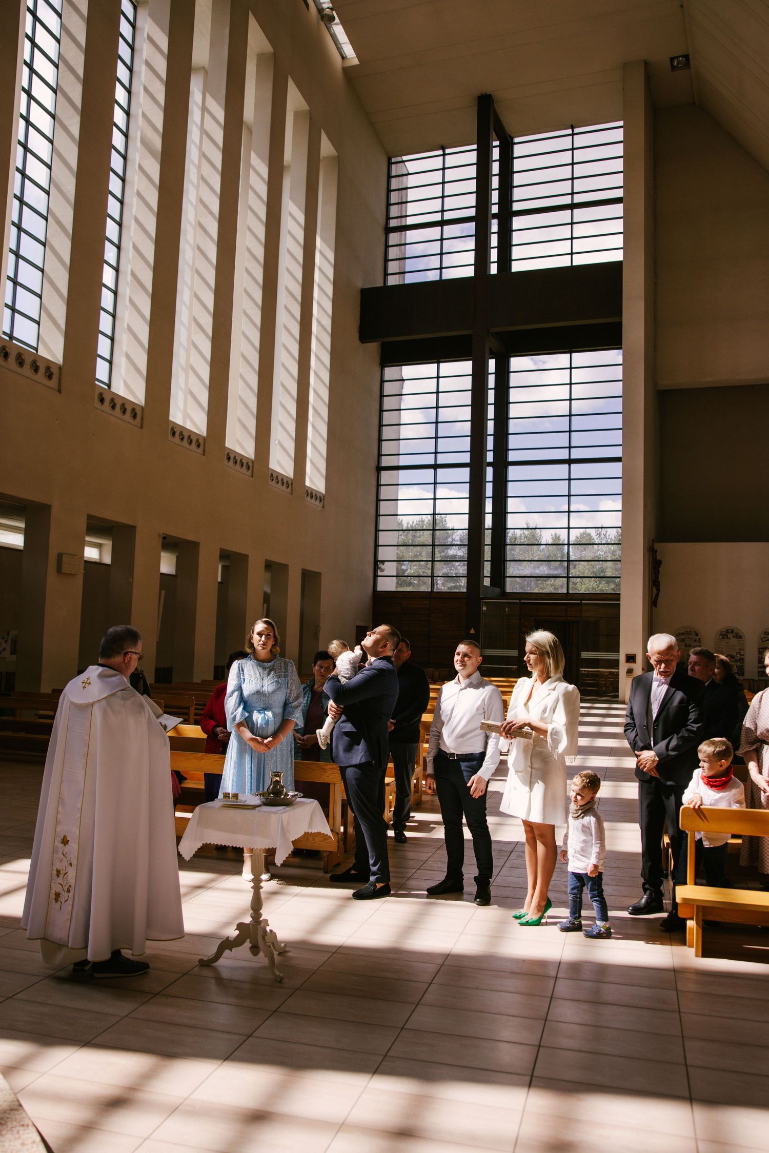 A religious ceremony taking place in a modern church interior with tall windows casting dramatic sunlight across the floor.