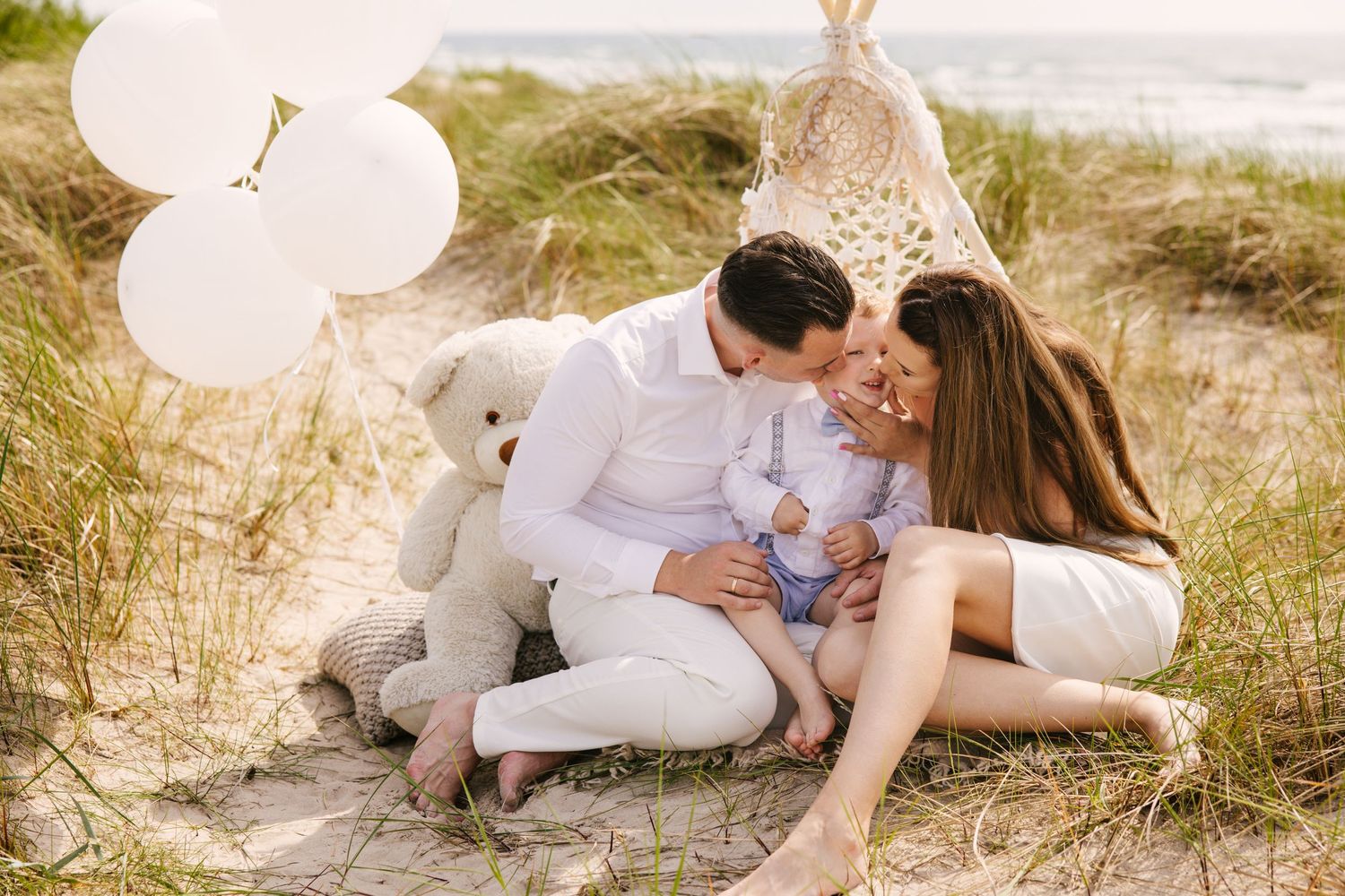 Family relaxing together on sandy dunes with white balloons during golden hour at the beach.