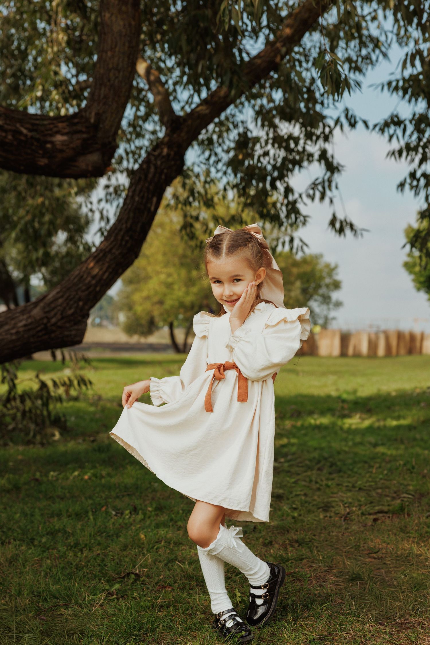 A child in a white dress with knee socks and black shoes twirls playfully under a large tree in a park setting.