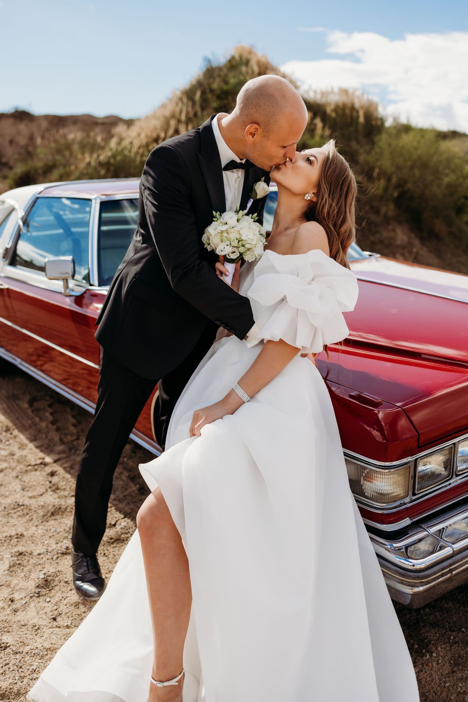 A romantic wedding photo with a couple kissing next to a vintage red car in a desert landscape.