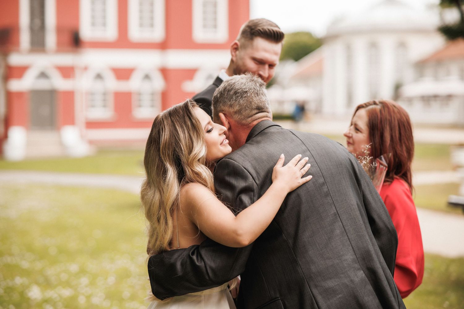 A group of people share an emotional embrace outdoors in front of a red building on a sunny day.