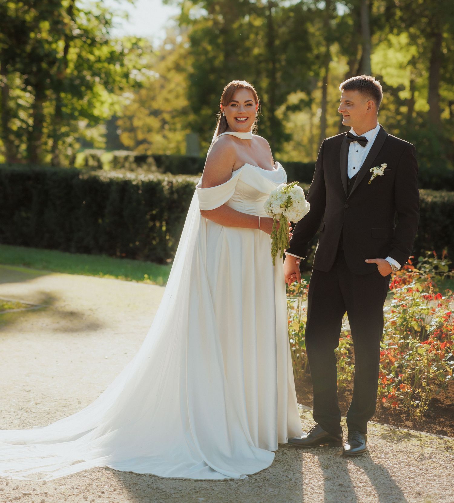Wedding couple walking along a sunlit garden path surrounded by autumn foliage.