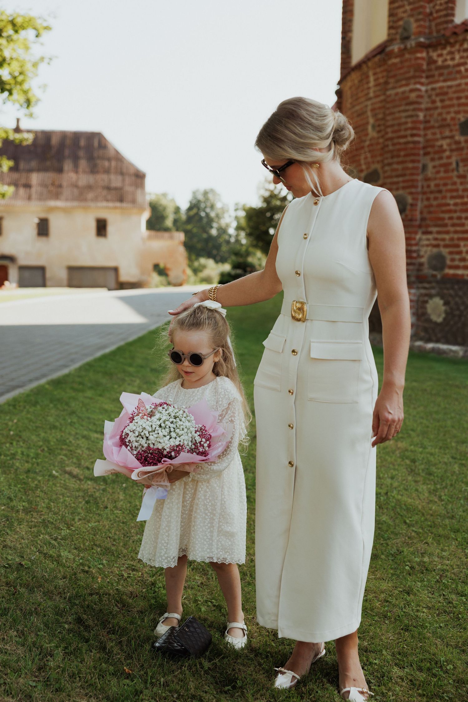 A woman in an elegant white dress stands with a young girl holding a pink floral bouquet on a sunny lawn.