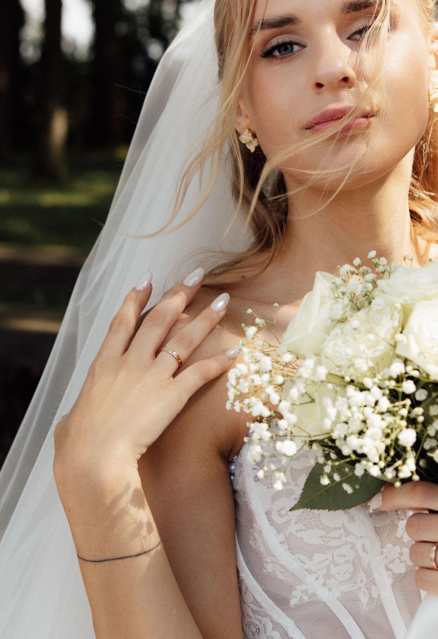 Elegant bride in white lace gown and veil holding white rose bouquet with baby's breath outdoors.