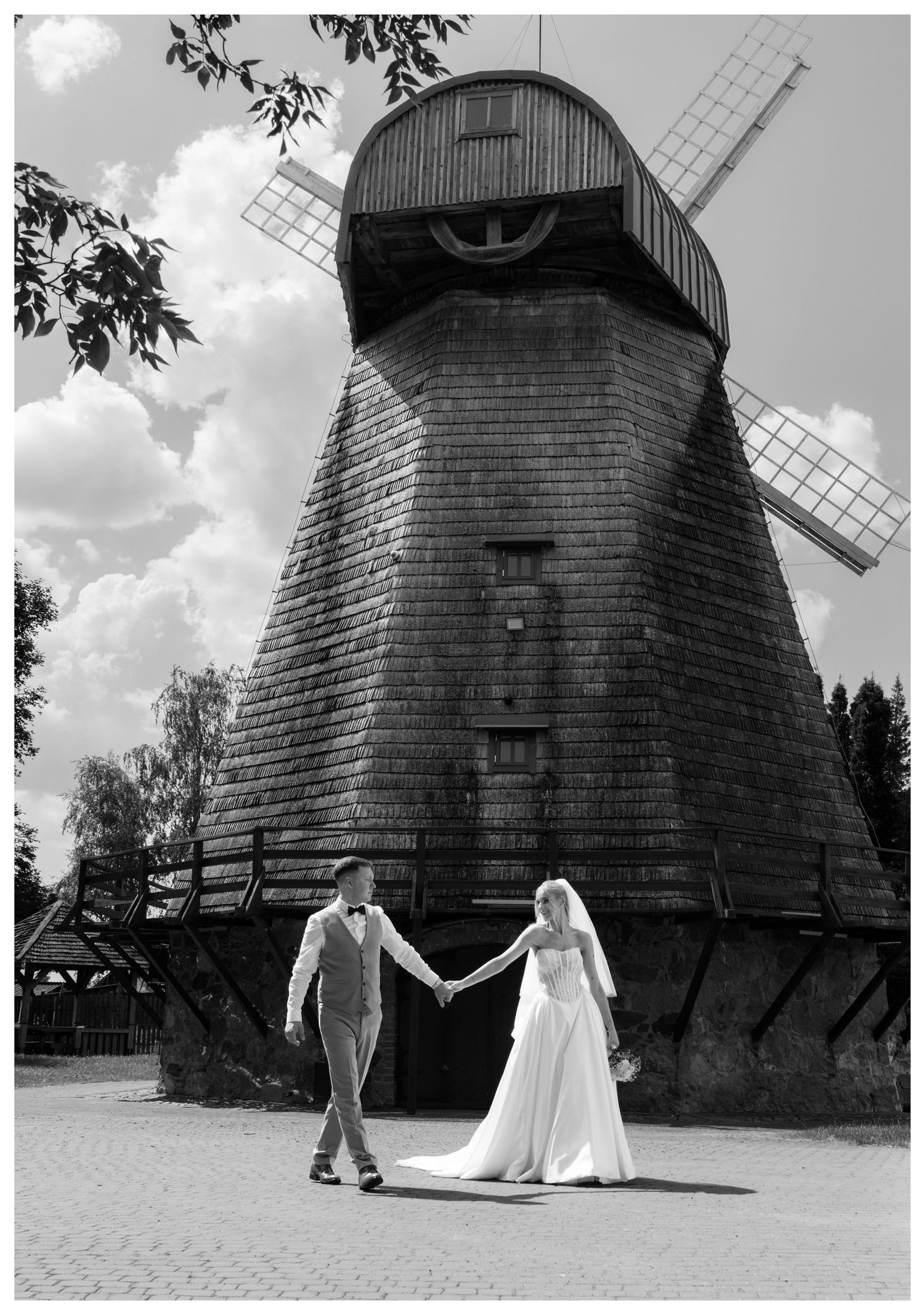Black and white photo of bride and groom holding hands in front of a large wooden windmill.