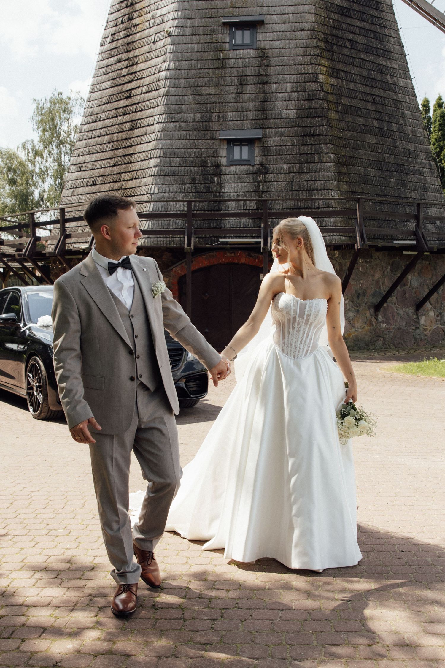 A bride and groom hold hands walking near a rustic wooden windmill on their wedding day.