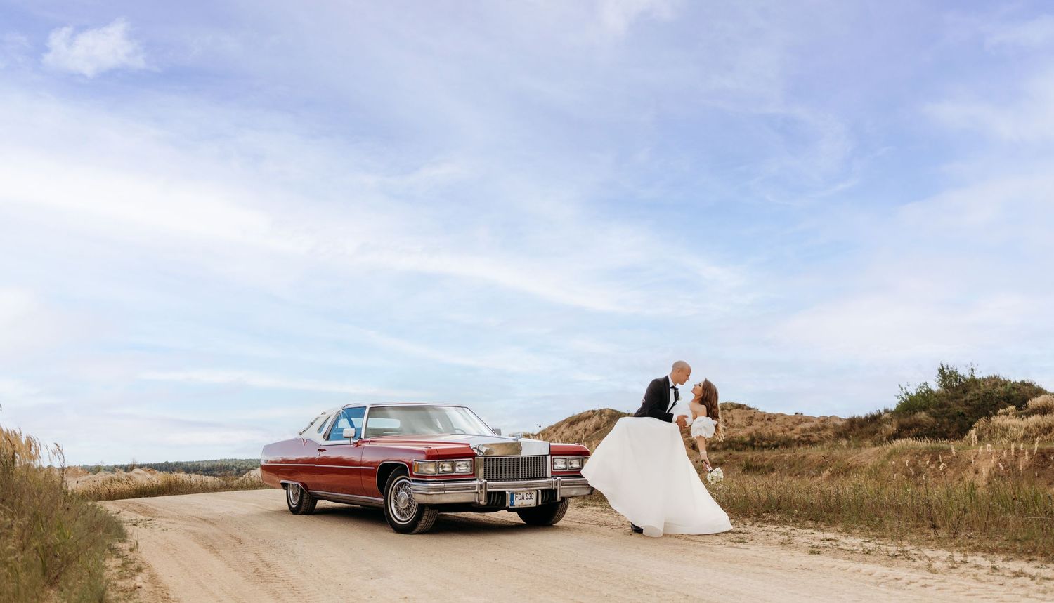 A classic silver car parked on a sandy beach with dunes and blue sky in the background.