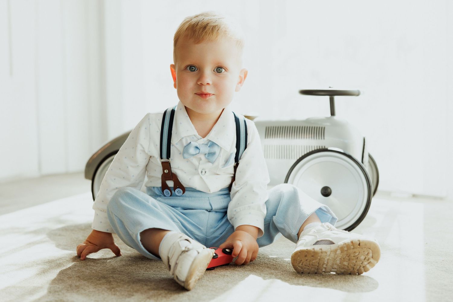 Small child in light blue shirt and suspenders lies on white floor while playing with toys.