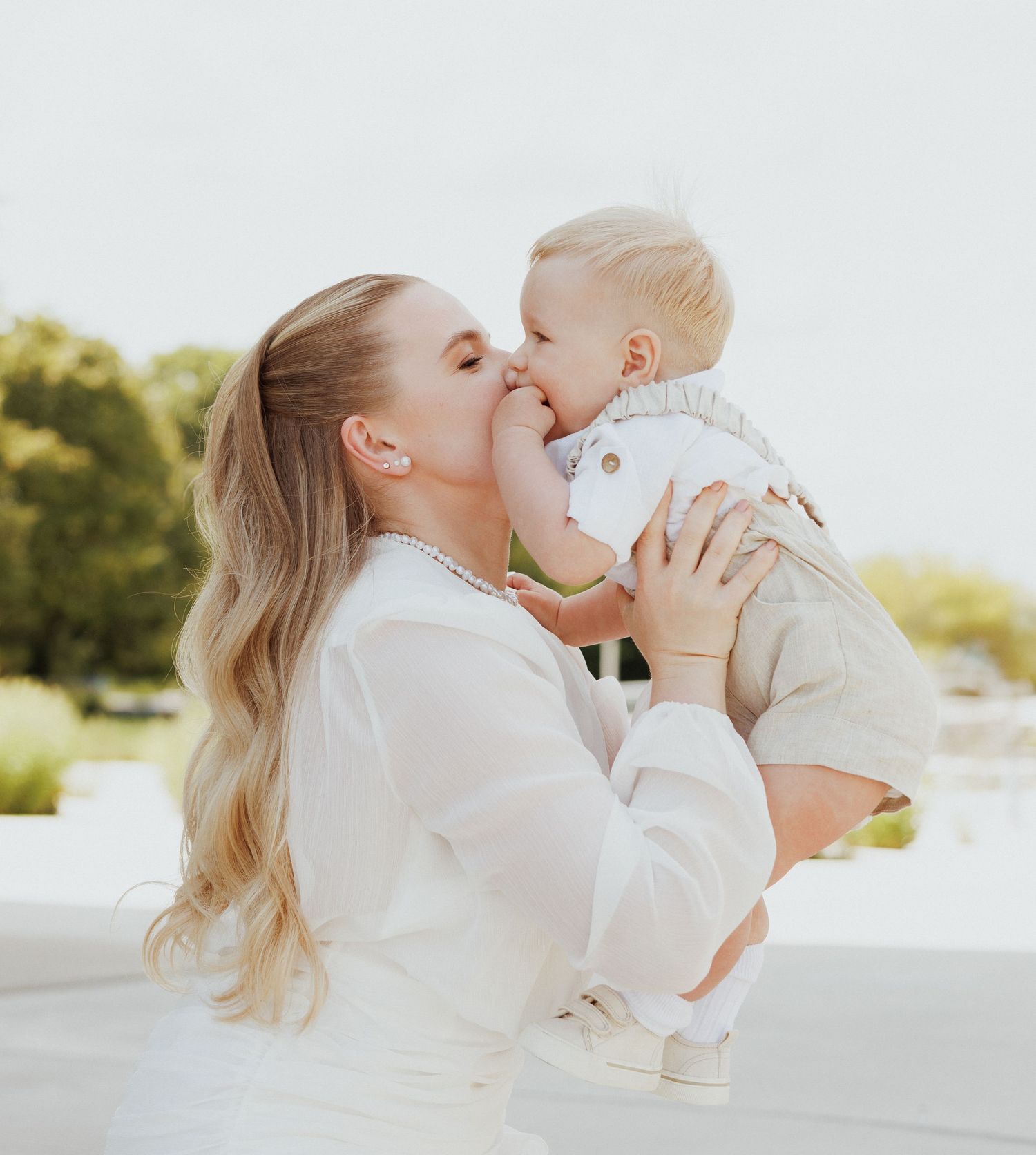 A tender moment between parent and baby in white outfits against a bright outdoor backdrop.
