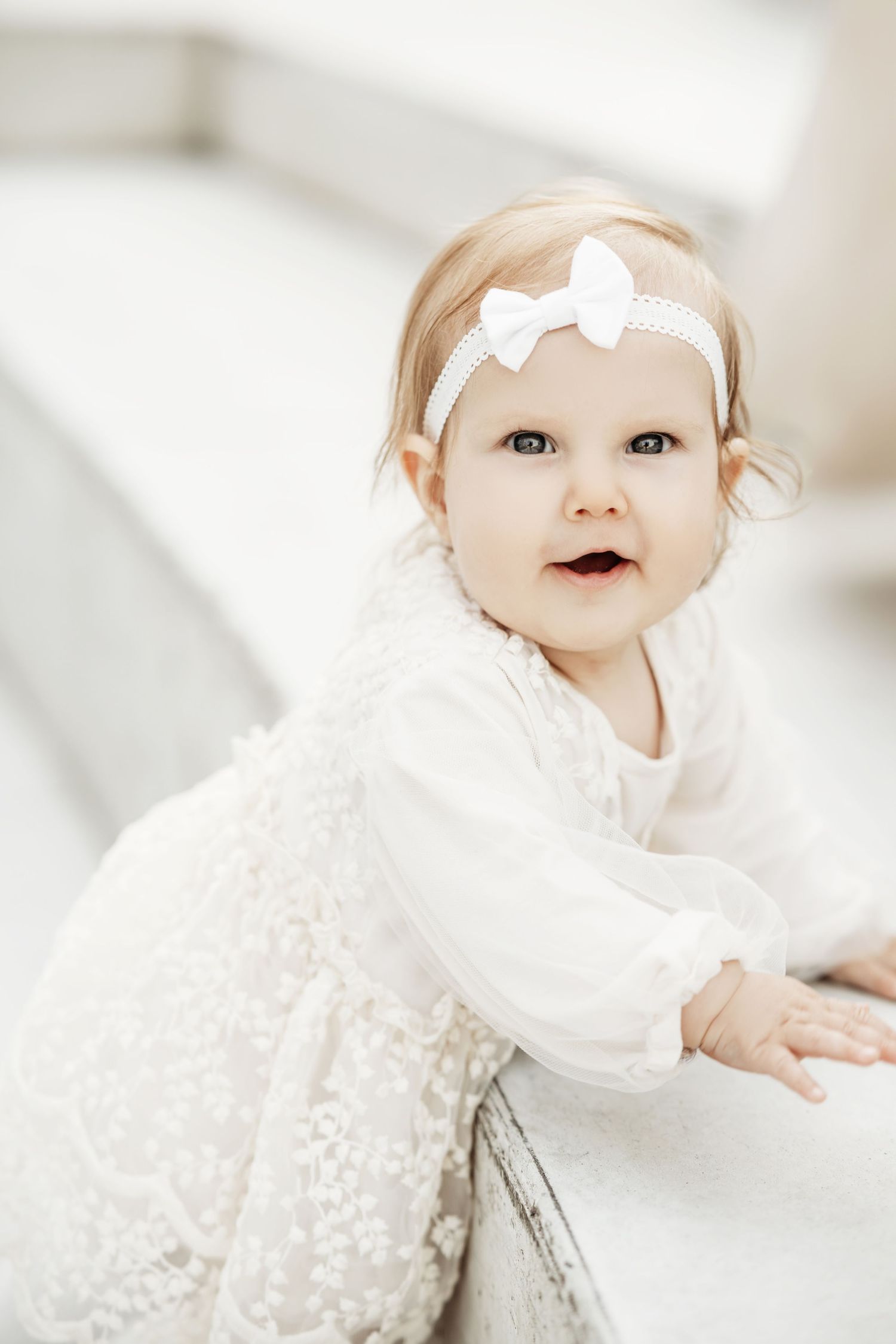 Baby wearing white lace dress and bow headband lying on soft white bedding in bright natural light.