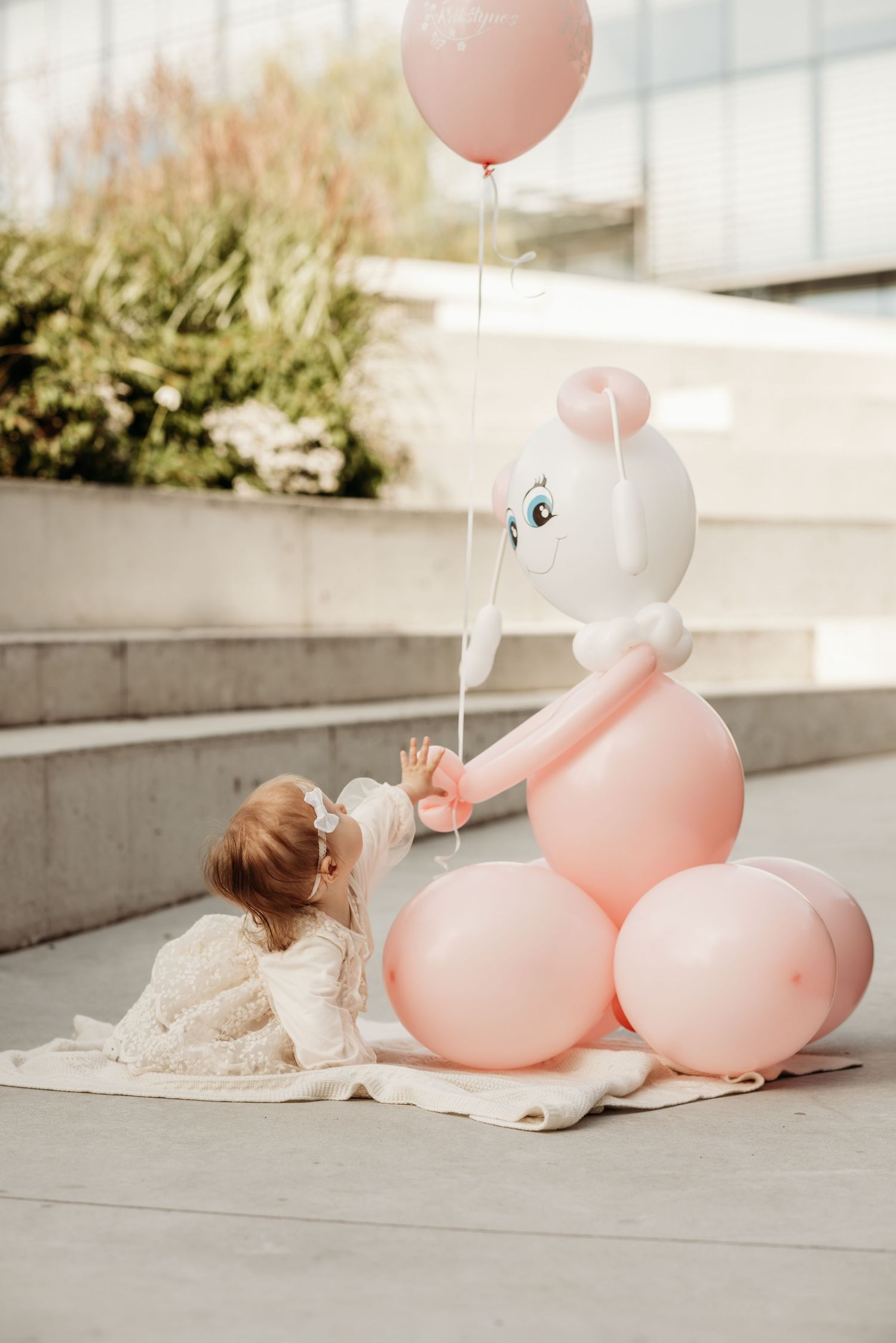 Pink balloons arranged beside small child in white dress laying on concrete steps outdoors.