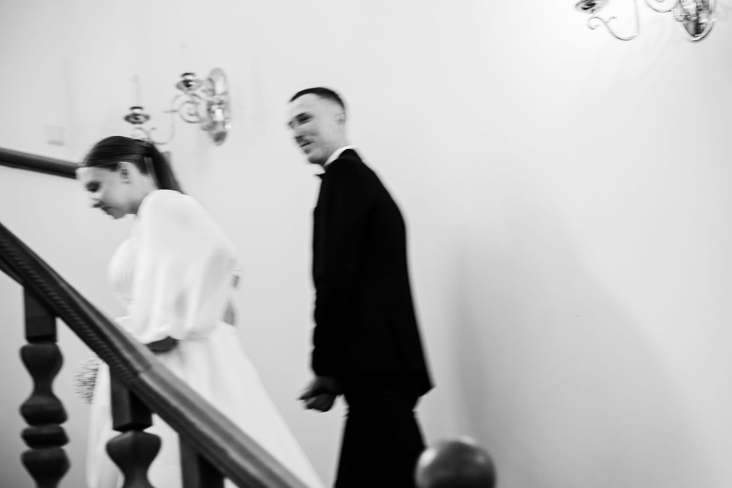 A artistic black and white photo of a couple descending a staircase with chandelier lighting.