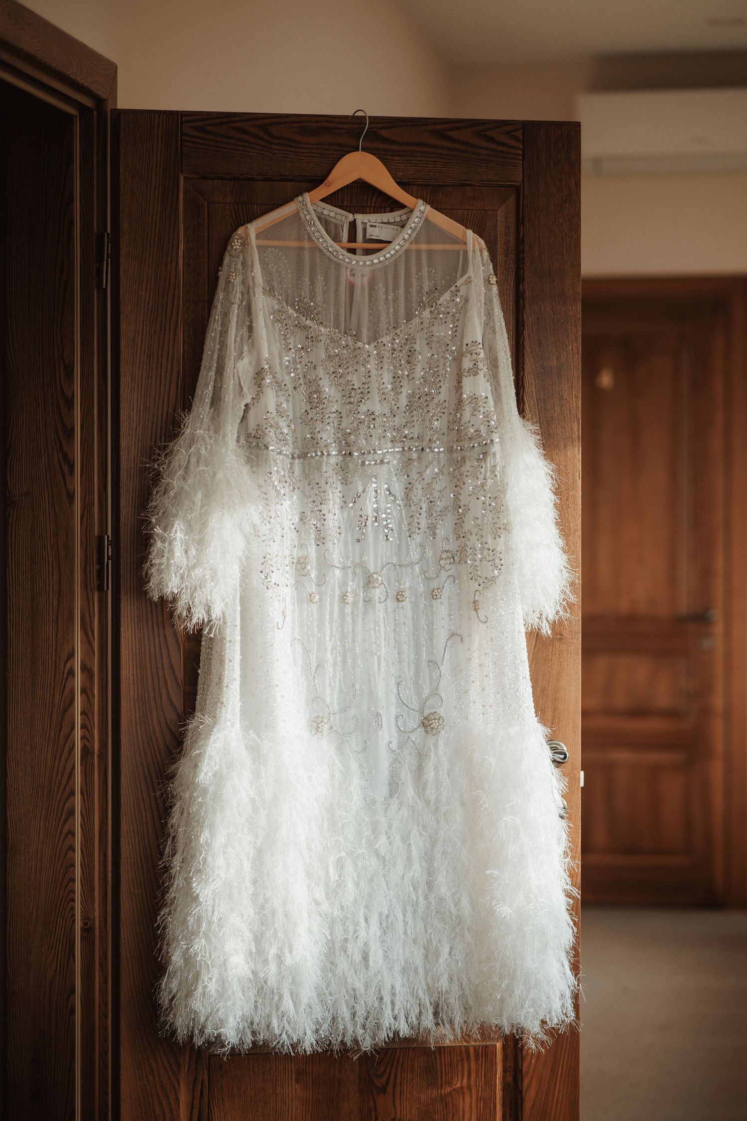 White feathered wedding dress hanging on wooden hanger against dark wall.