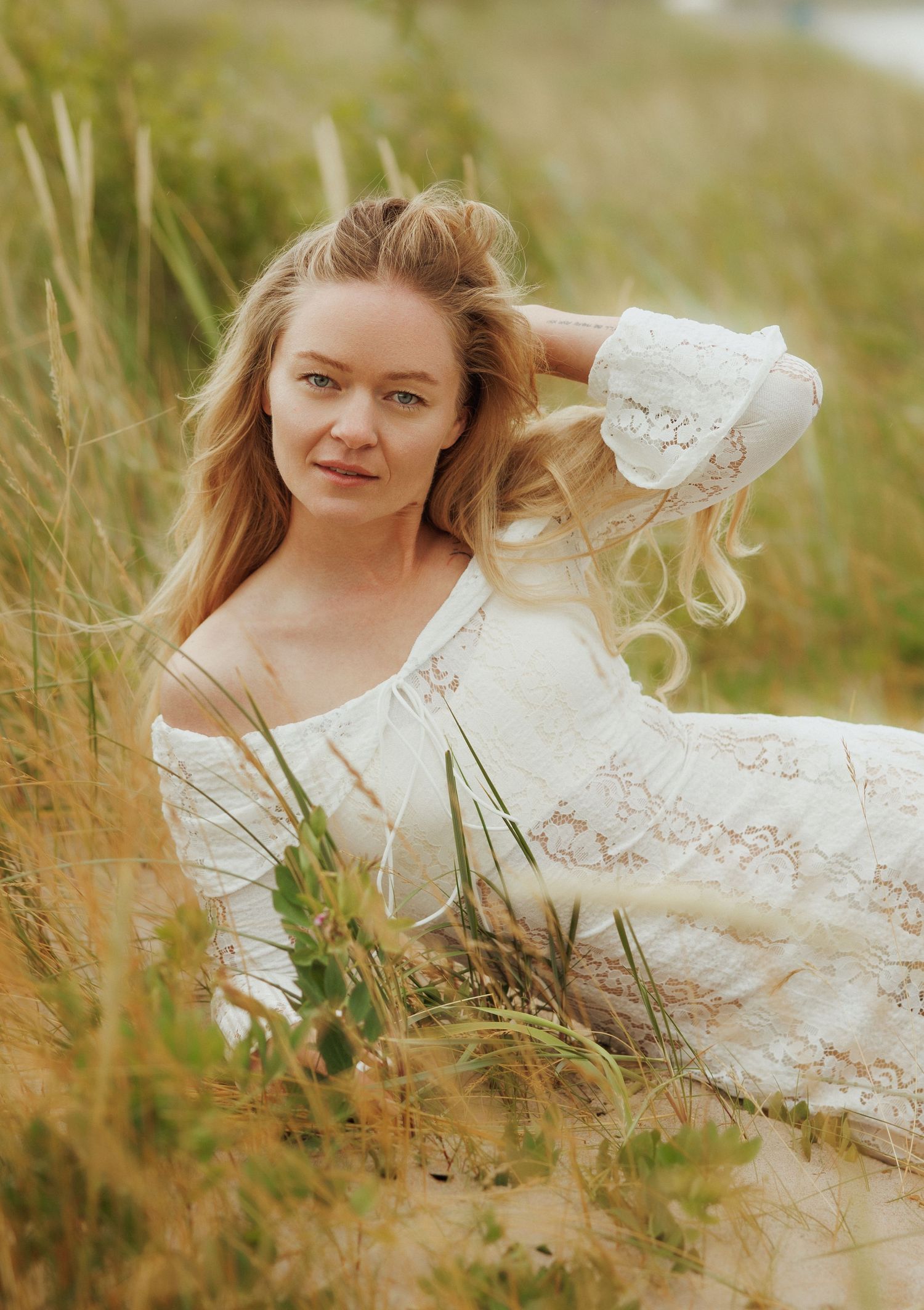 Blonde woman in white lace off-shoulder dress sitting among tall grass dunes, posing with hand in hair.