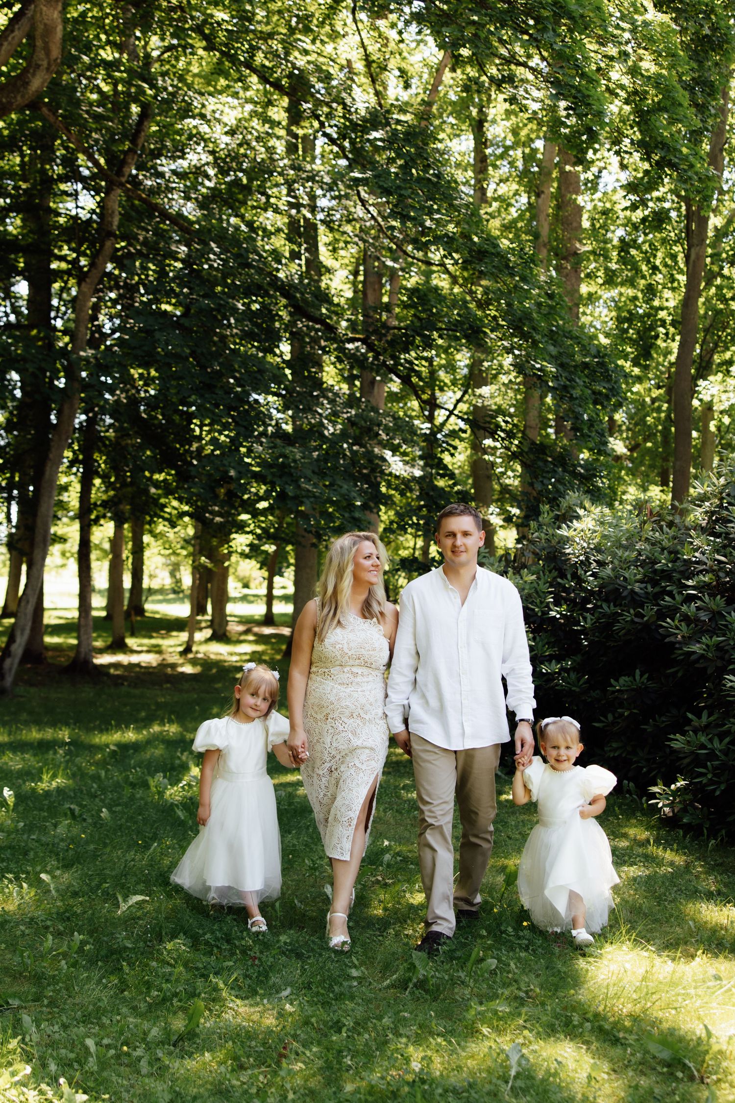 Happy family of four walking in a sunny green park, parents and two young girls in white dresses holding hands.