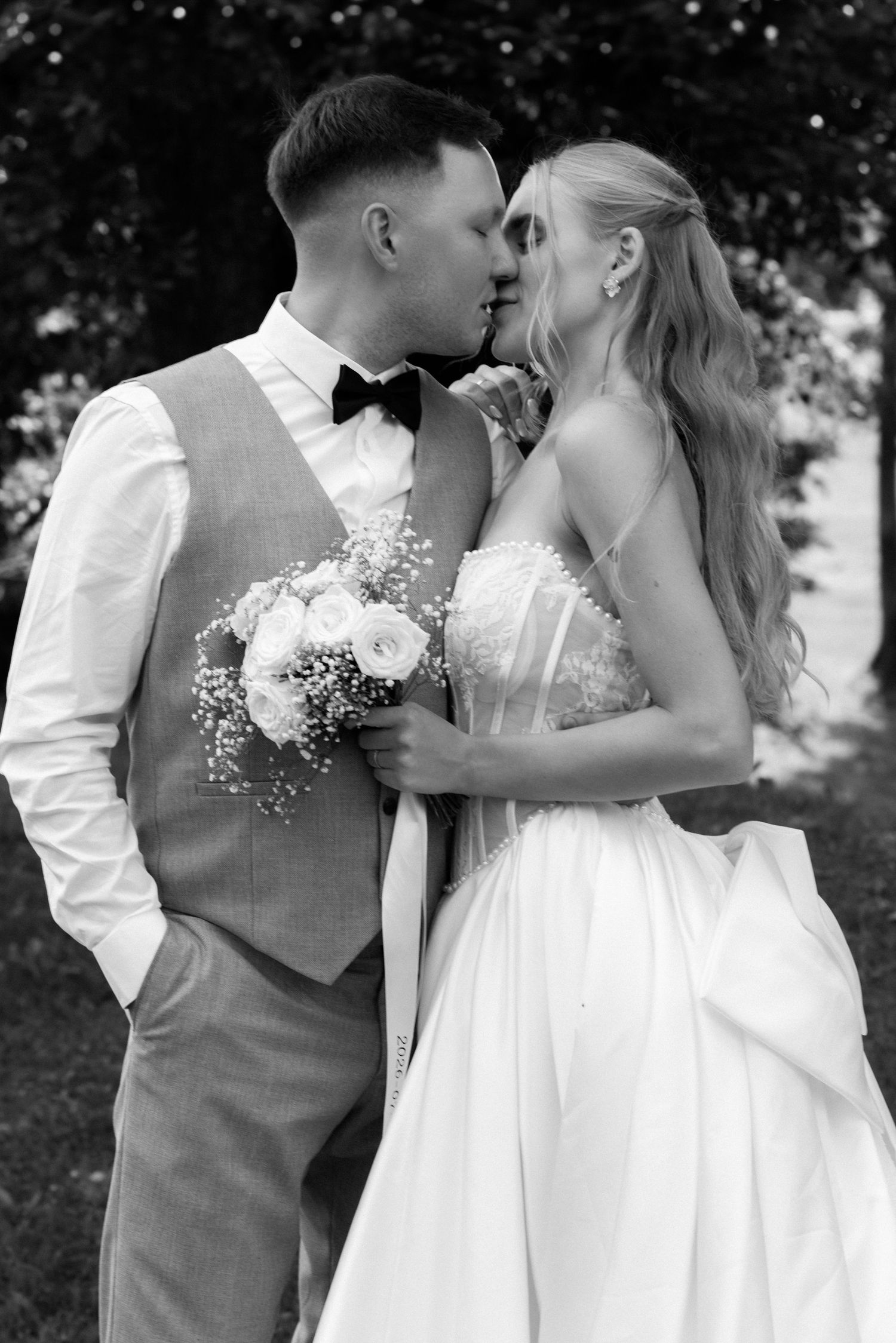 Black and white photo of a bride and groom sharing a kiss outdoors, bride holding a bouquet of white roses.