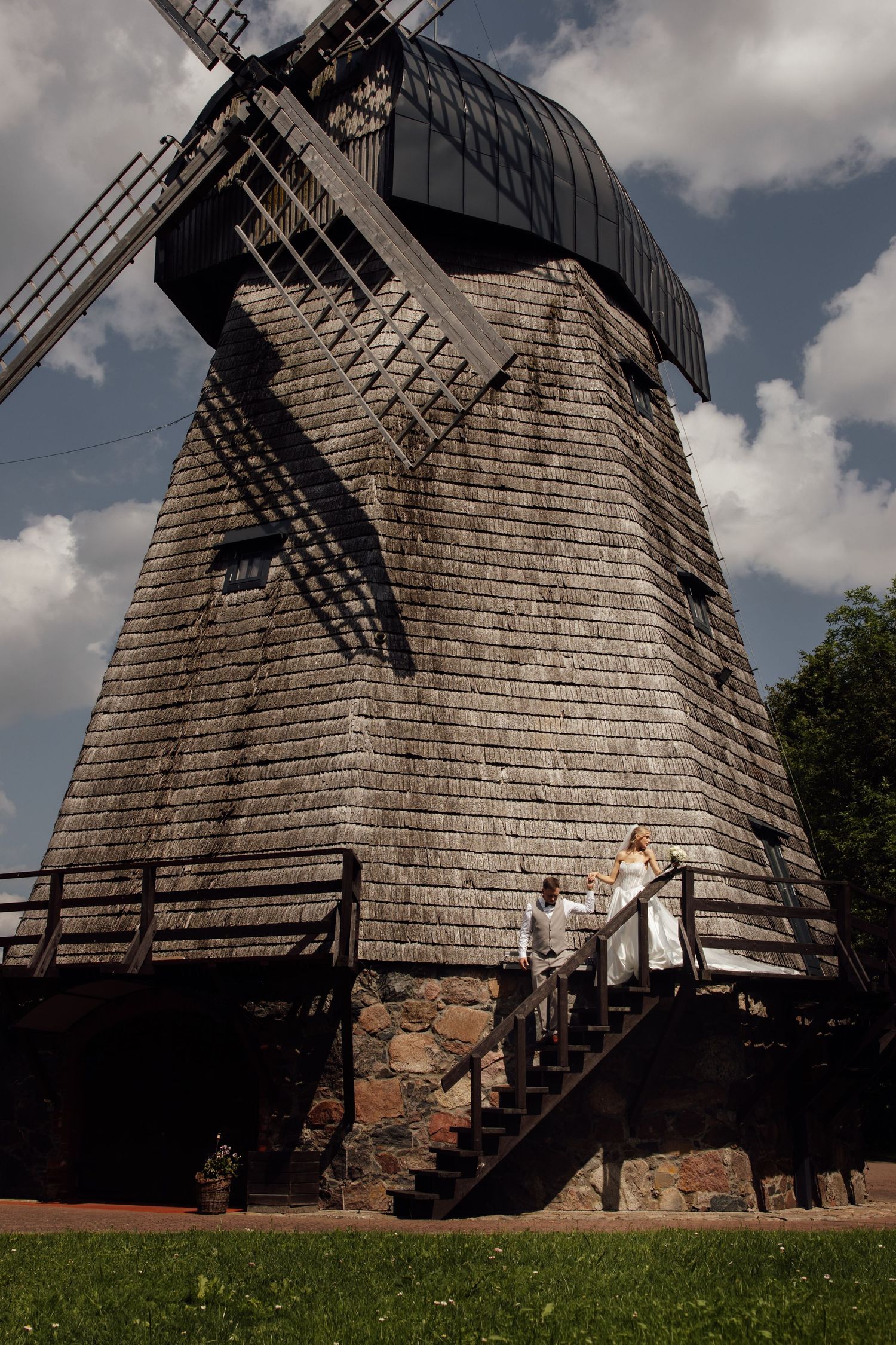 Bride and groom standing on wooden stairs of a historic wooden windmill under a cloudy sky.