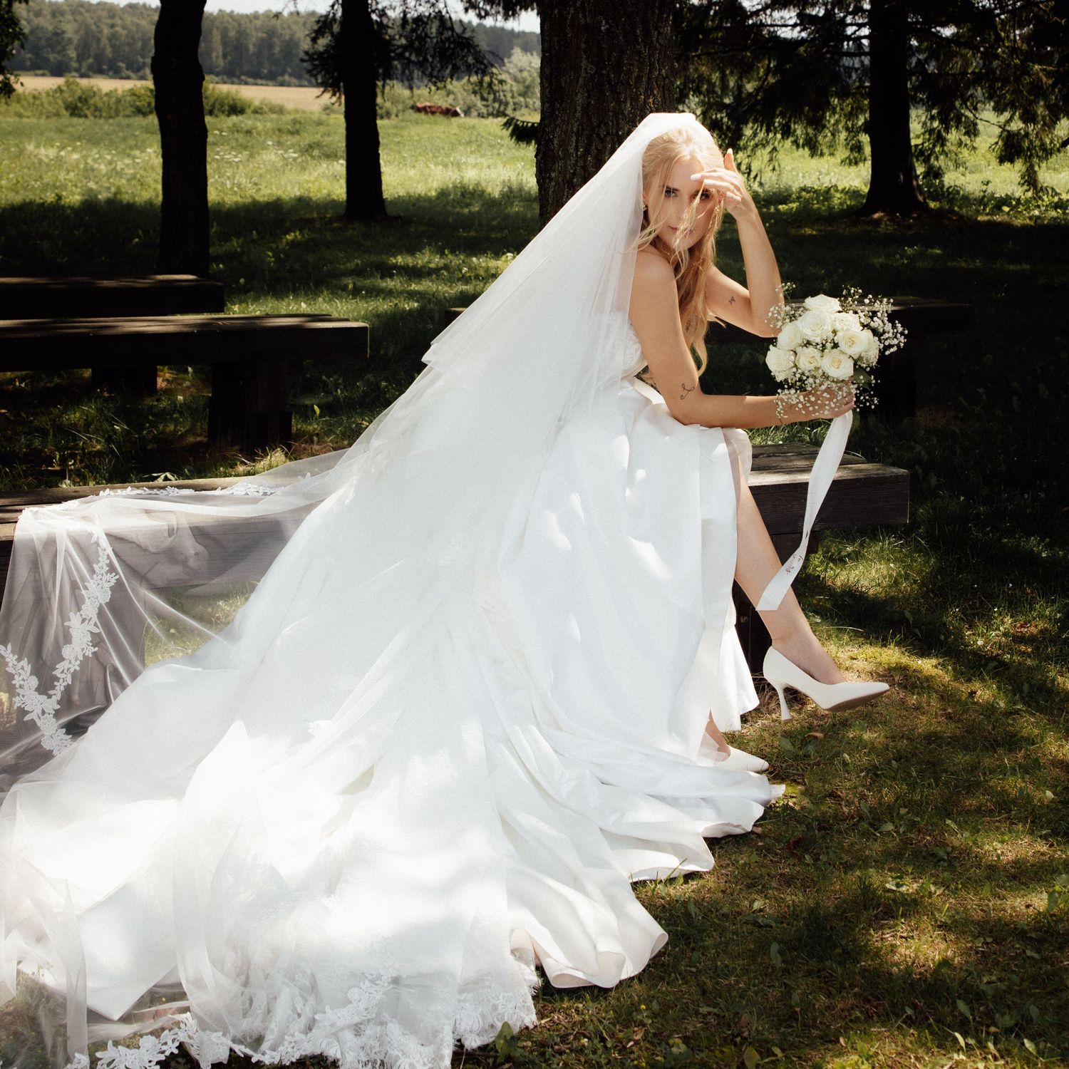 Bride in white wedding gown with long lace veil holding bouquet, seated outdoors in lush green park setting.