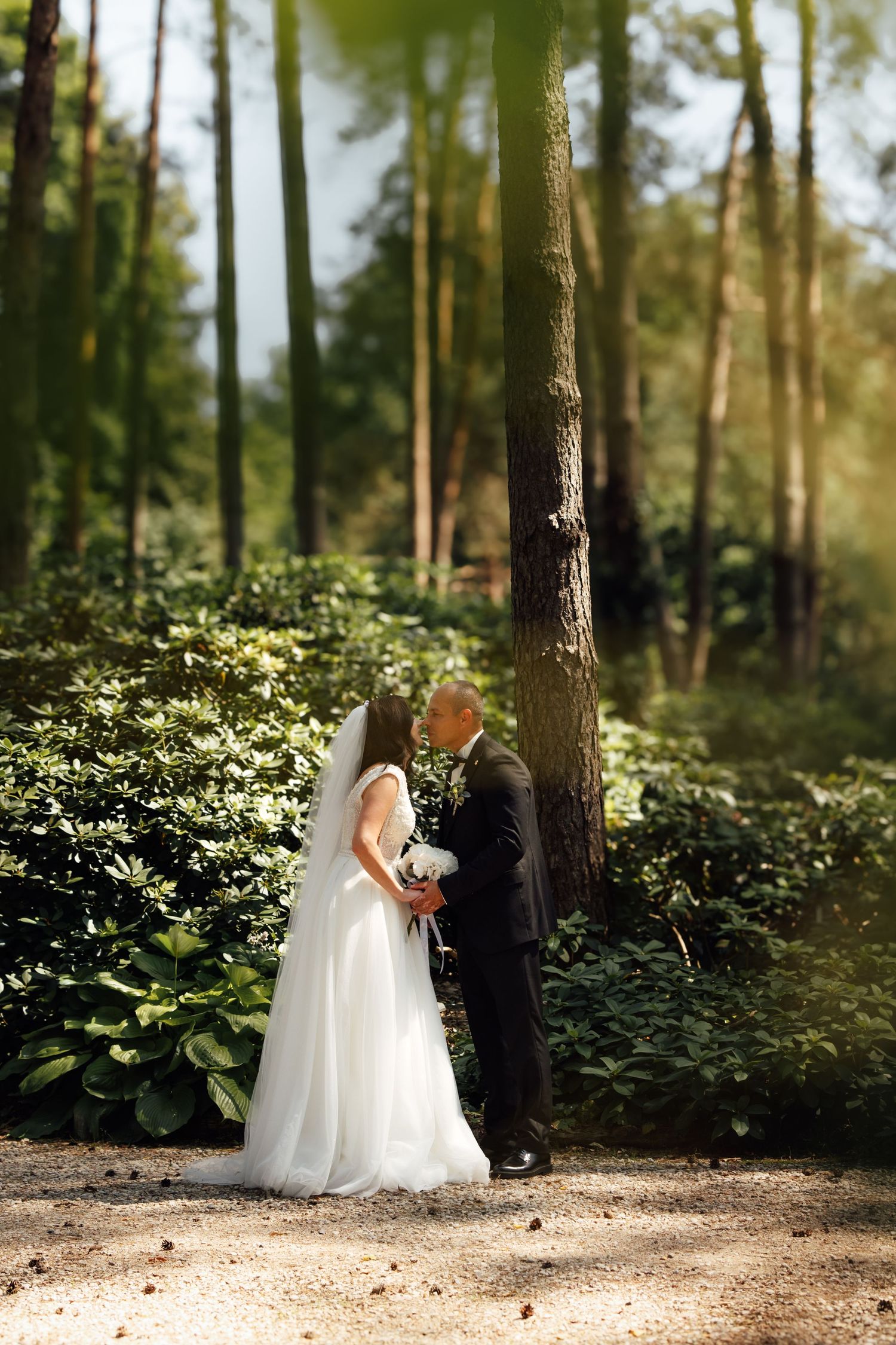 Bride and groom share a romantic moment in a lush green forest, bride wearing white gown with veil holding bouquet.