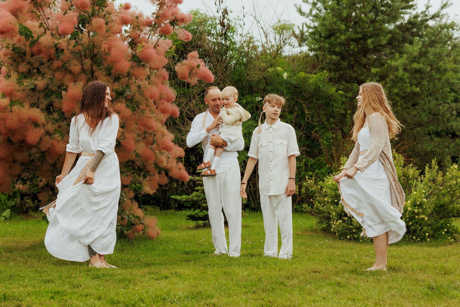 Family dressed in white outfits posing together in a lush garden with pink flowering trees.
