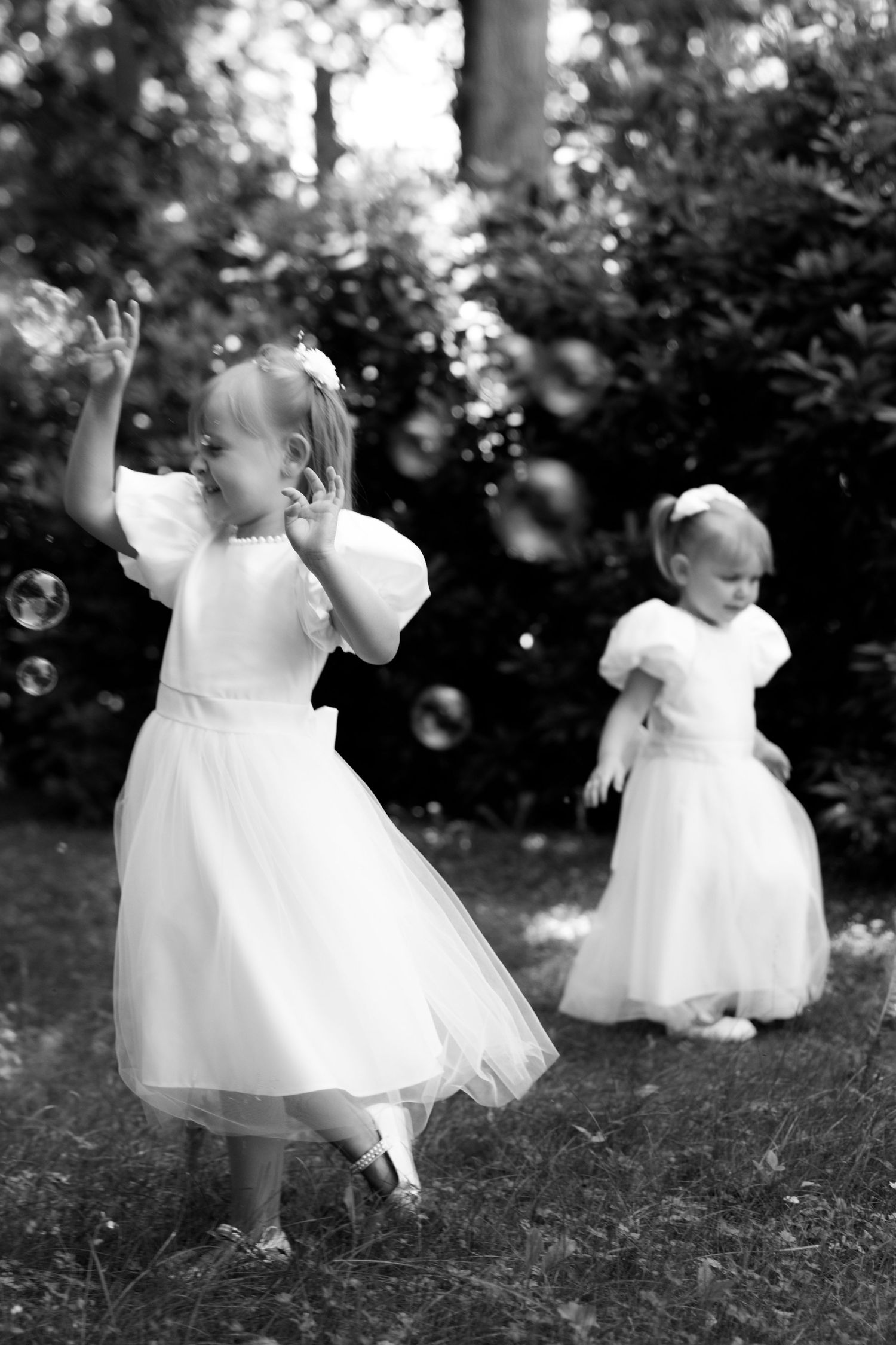 Two little girls in white dresses playing with bubbles outdoors in black and white photo.