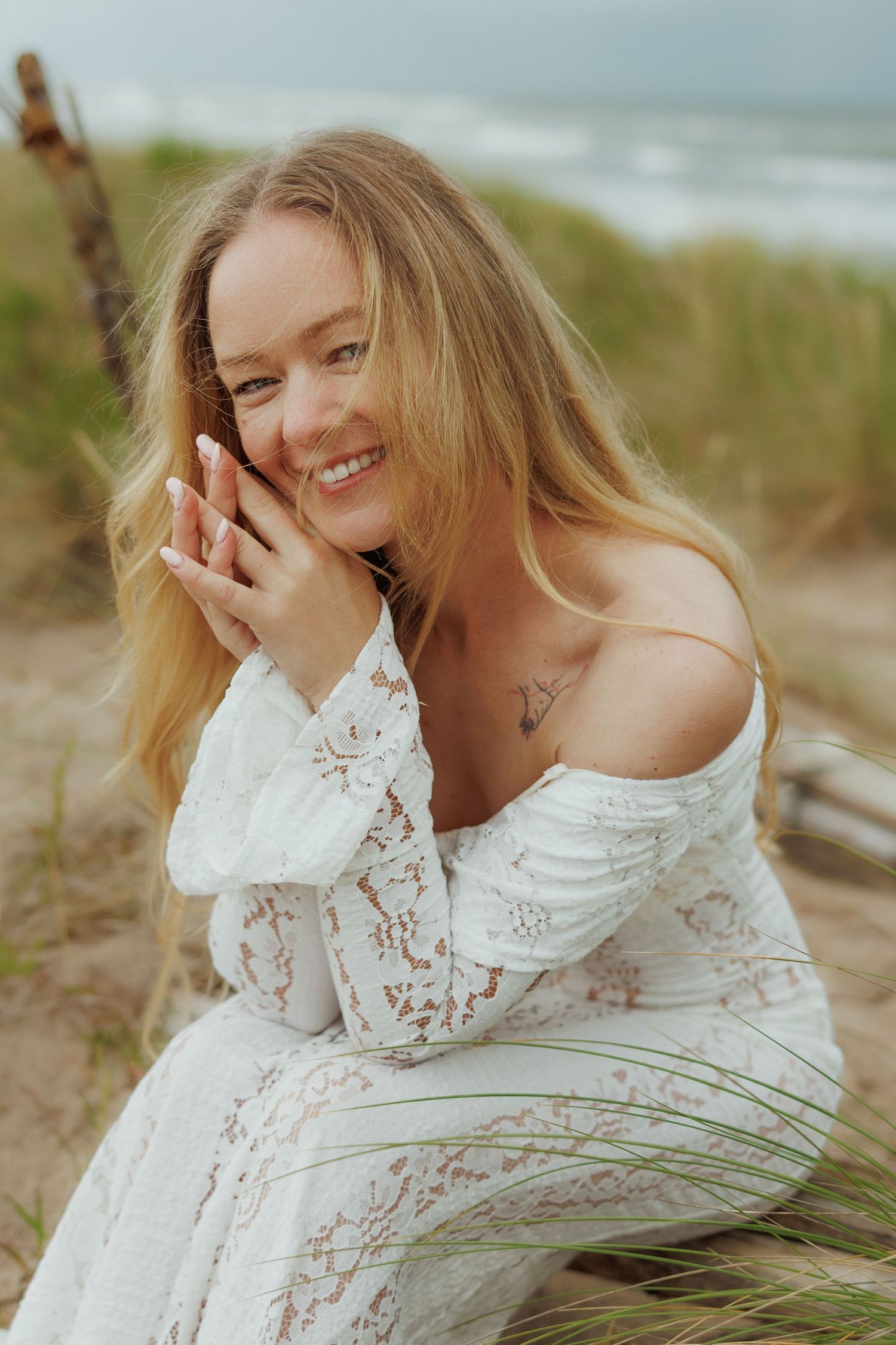Smiling blonde woman in white off-shoulder lace dress sitting on beach dunes with ocean in background.