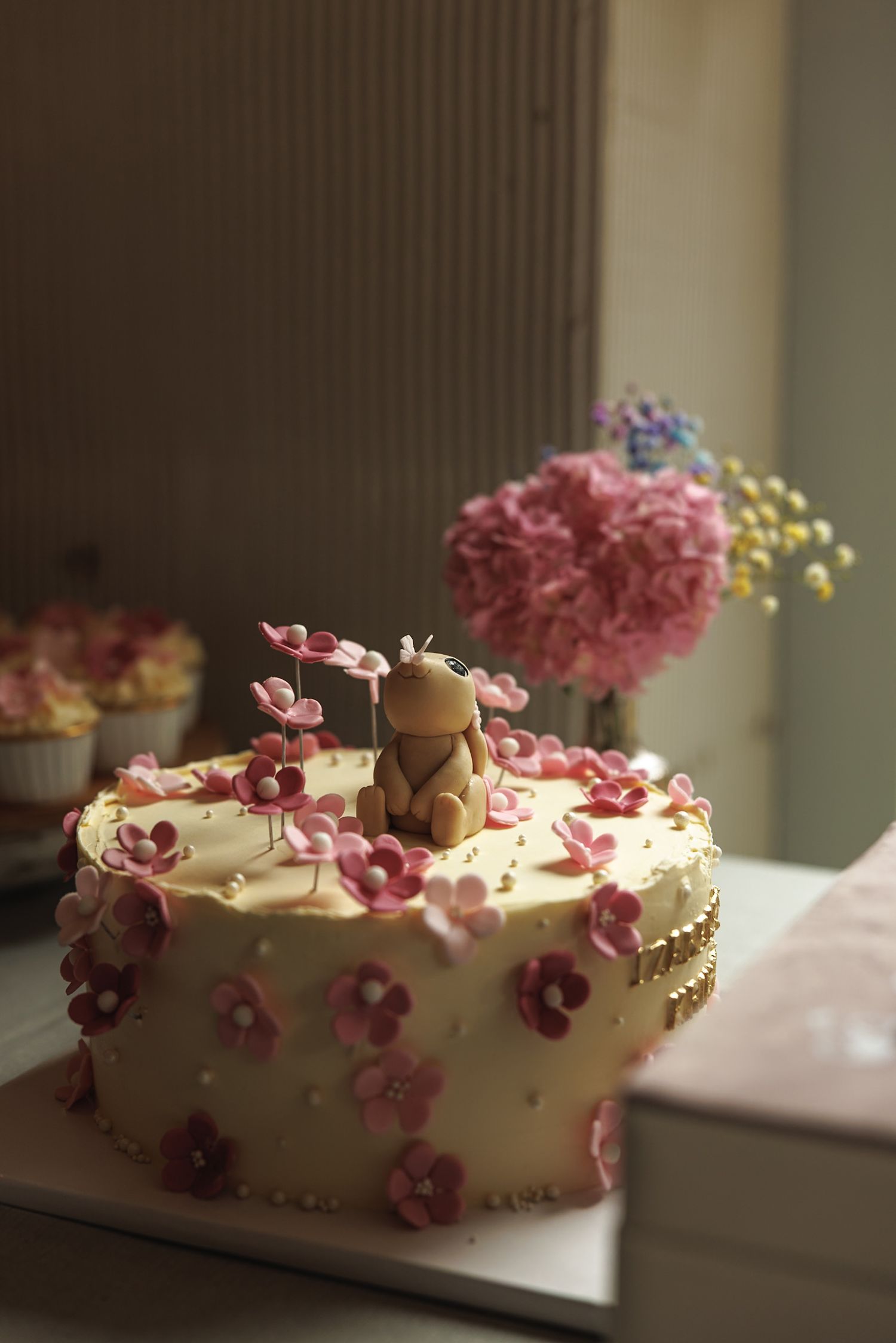 White celebration cake decorated with delicate pink sugar flowers and a teddy bear topper.