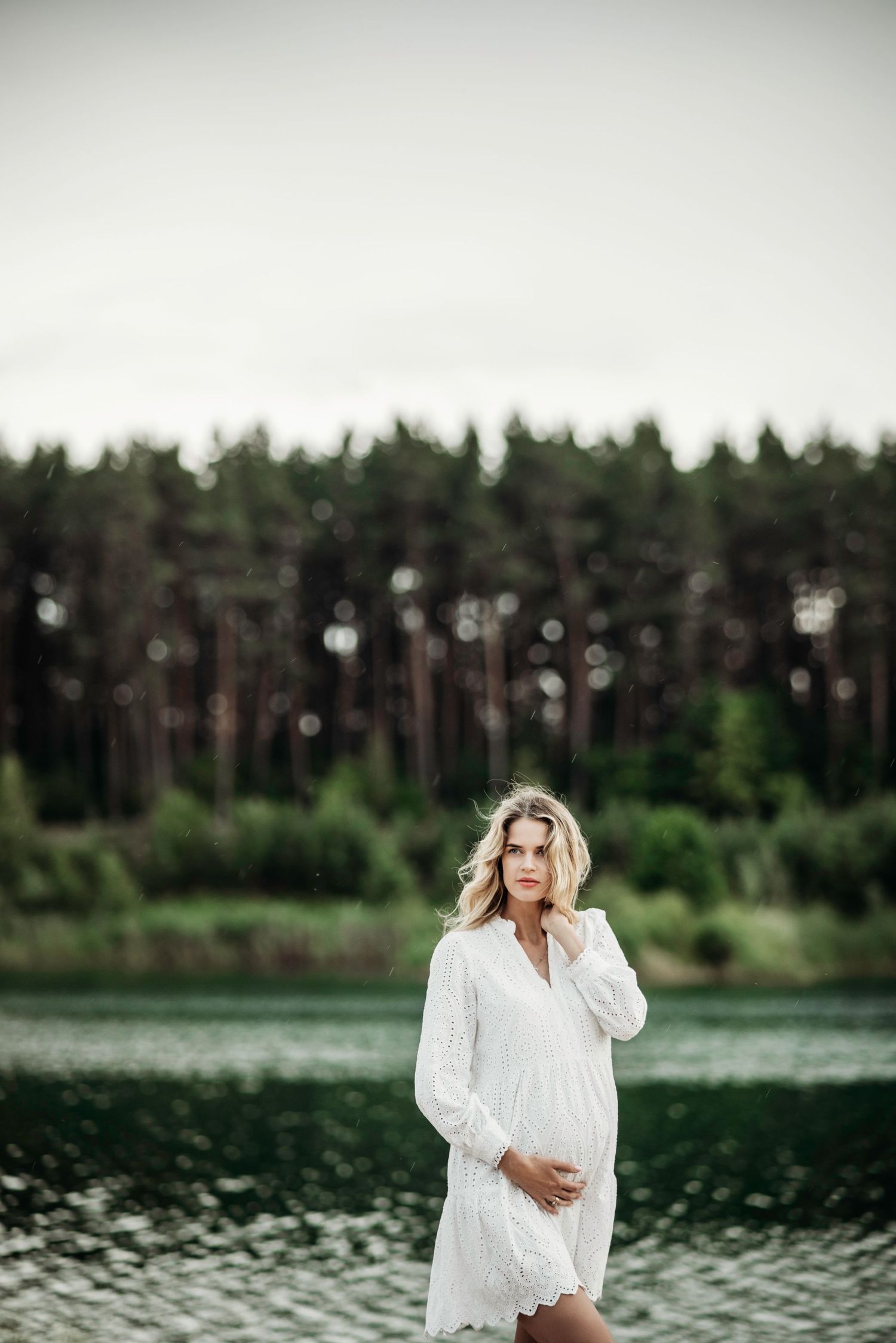 Serene maternity portraits by a lake with pine trees in the background.