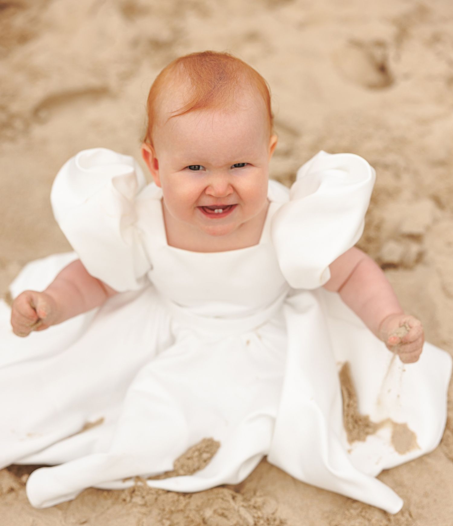 Baby wearing white baptismal dress lying on sandy beach background.