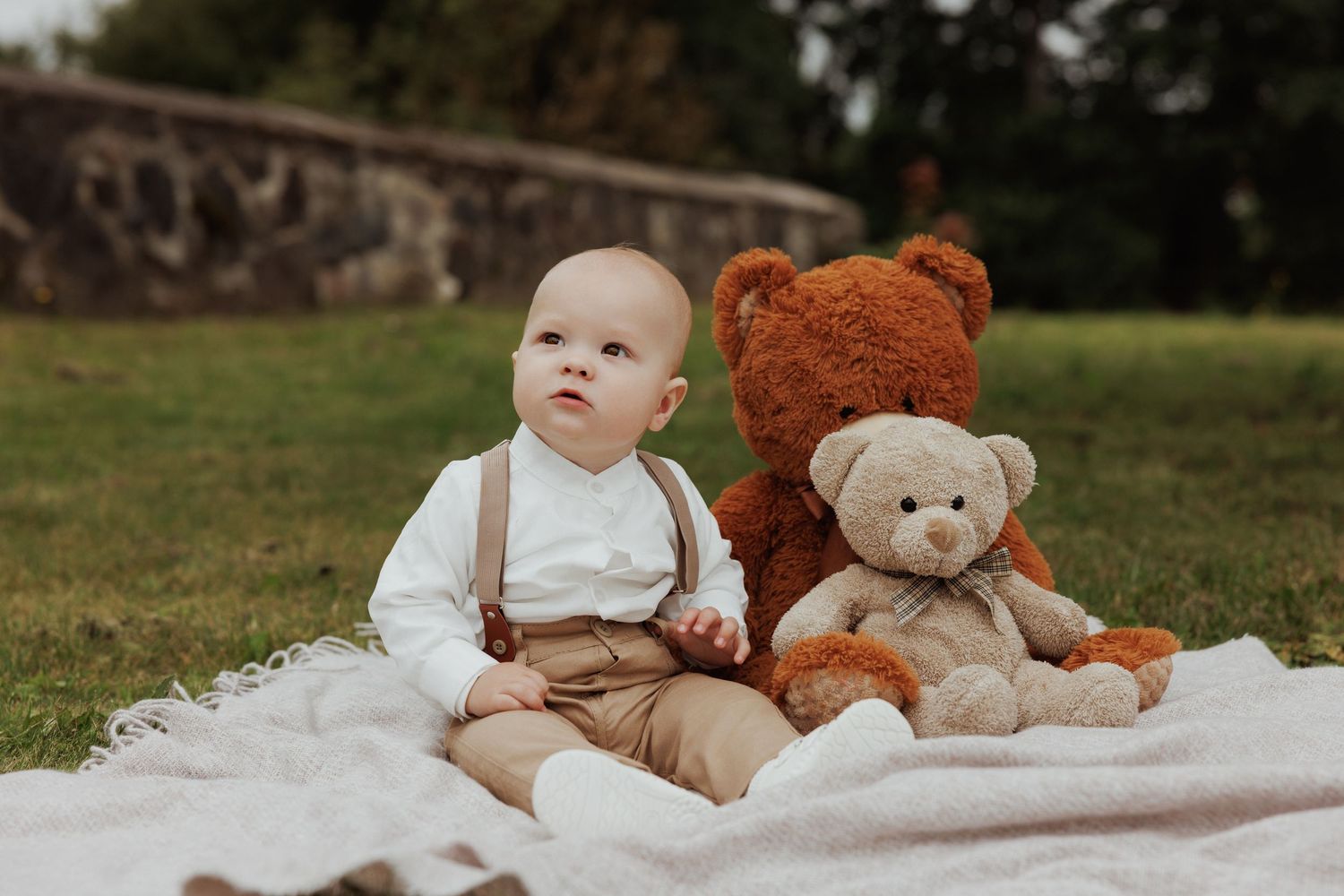 A baby in vintage-style suspenders sits on a blanket outdoors with two plush teddy bears.