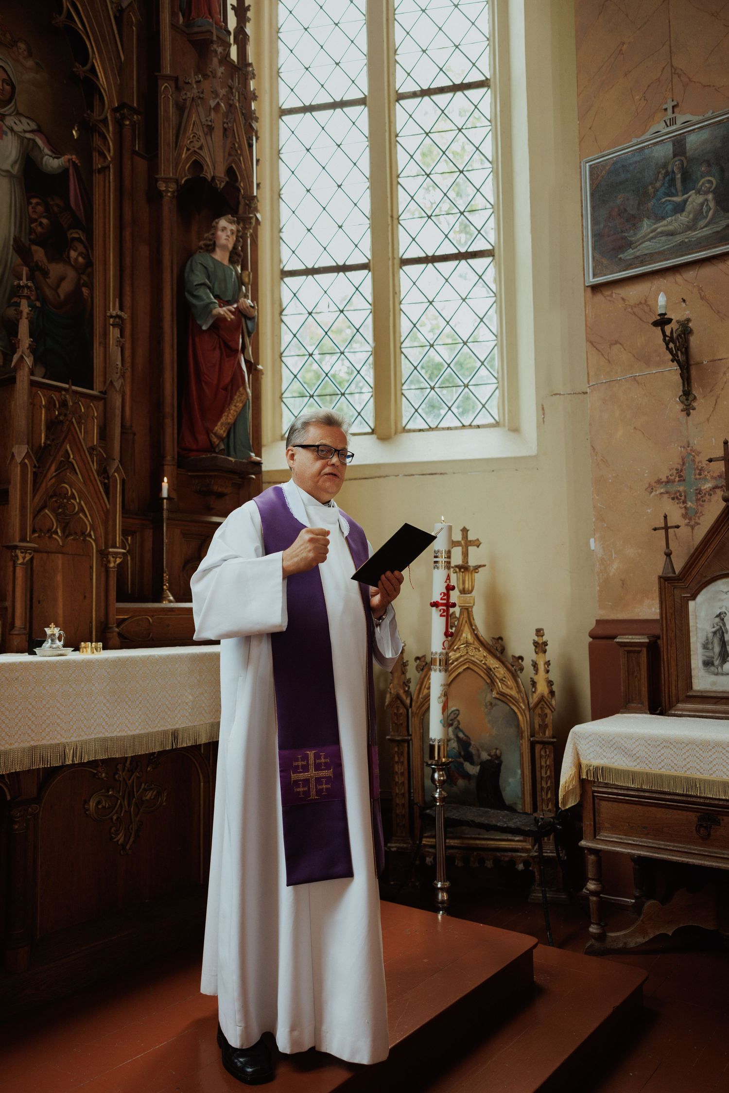 Priest in white robes and purple stole conducts a religious ceremony in a traditional church setting.