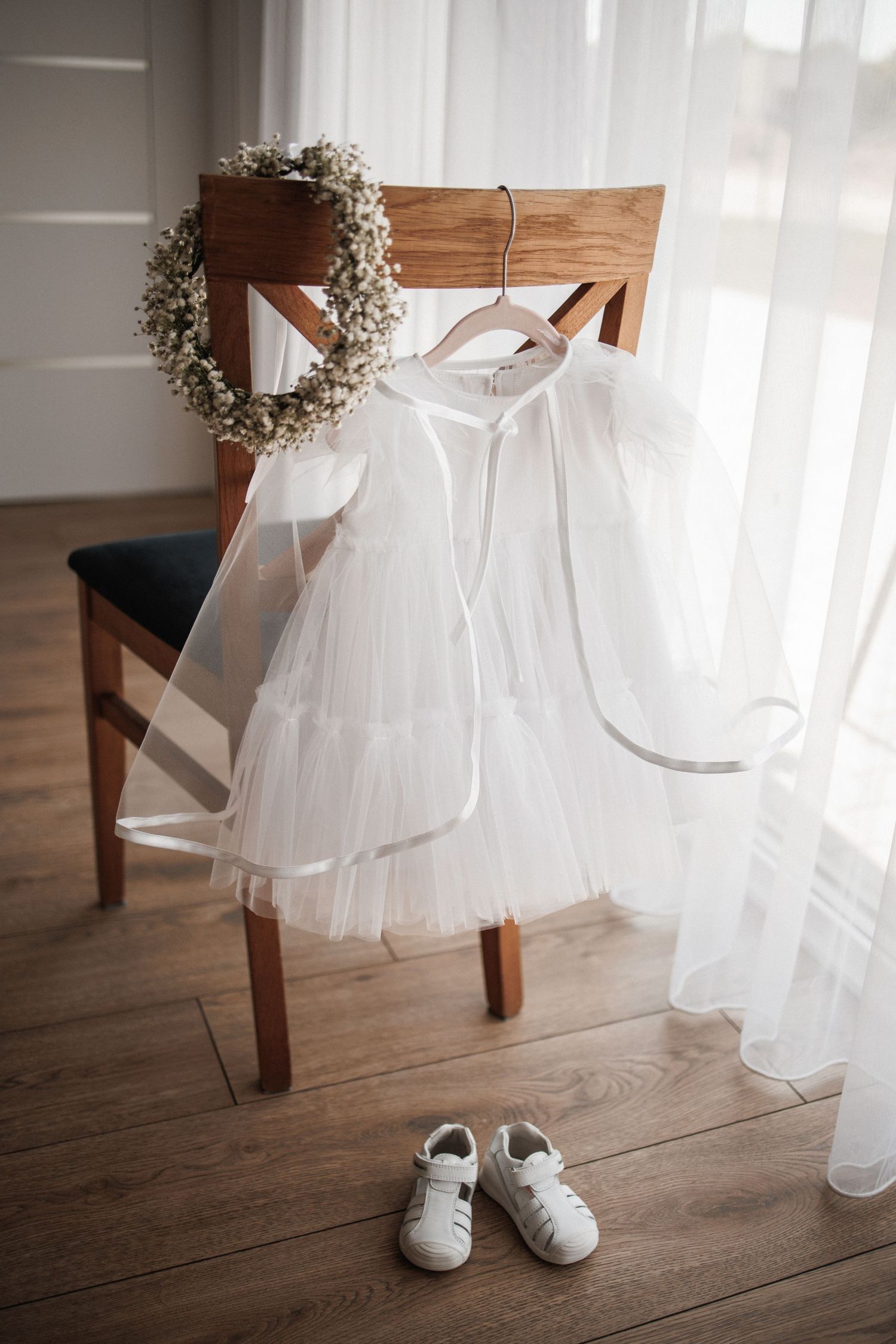 A white tulle flower girl dress hangs on a wooden chair next to small white shoes on hardwood flooring.