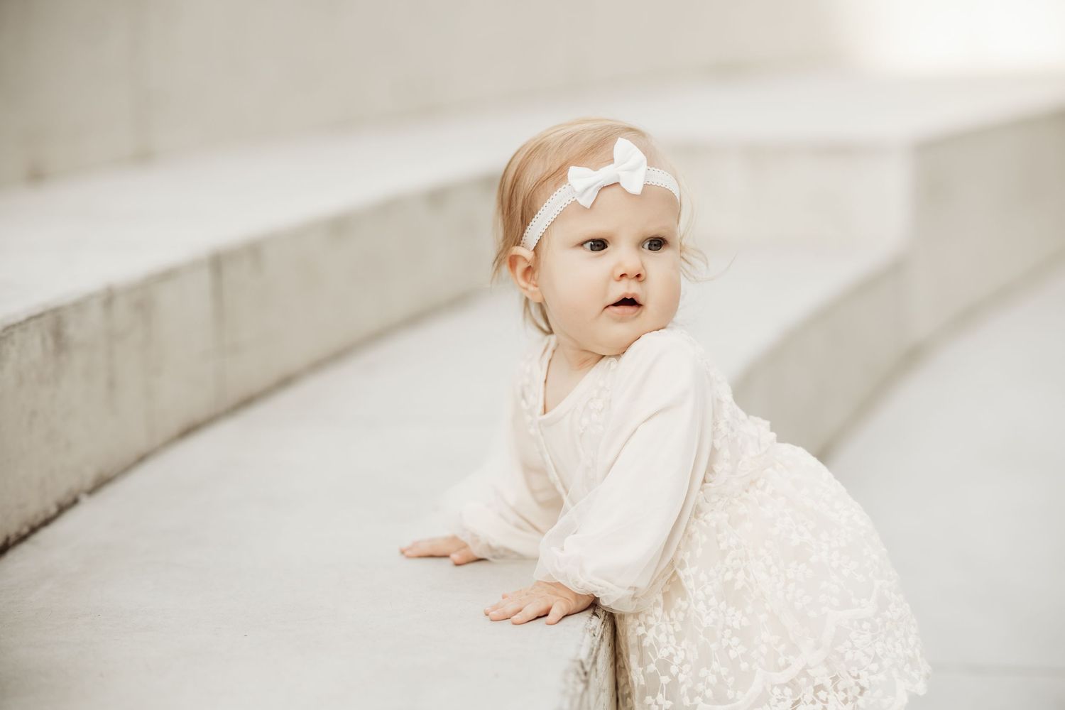 A baby in a white dress sits on a light colored floor and looks up with curiosity.