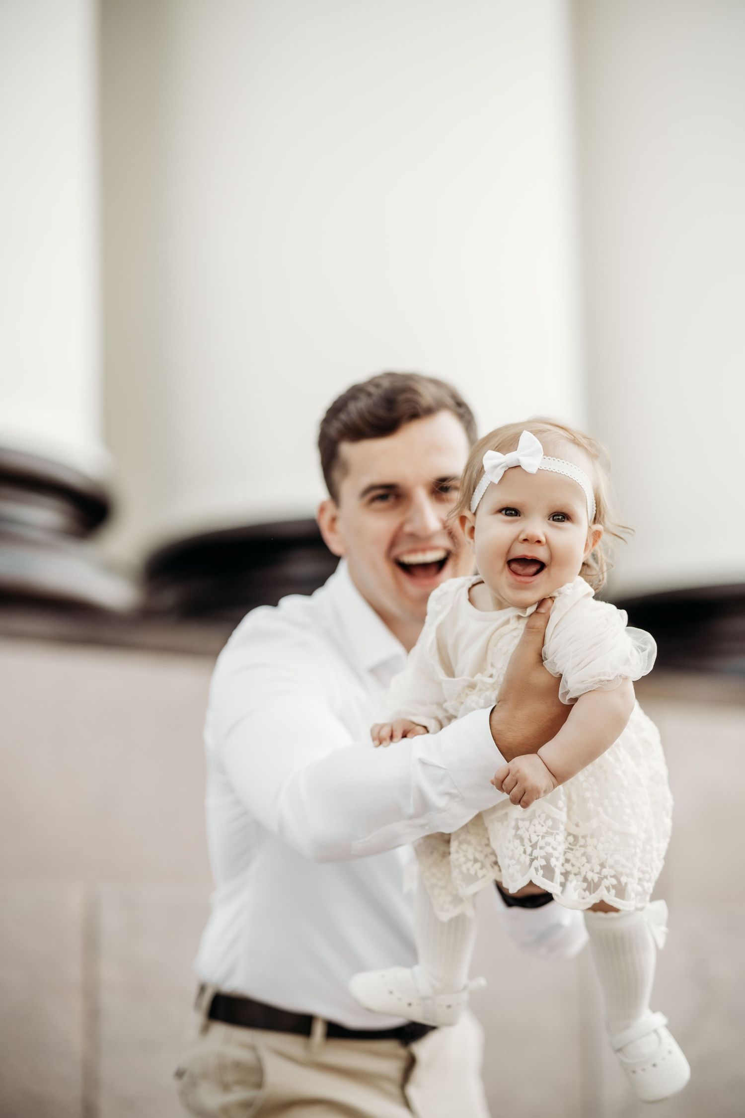 A tender moment with a baby in white lace dress being held up against a light background.