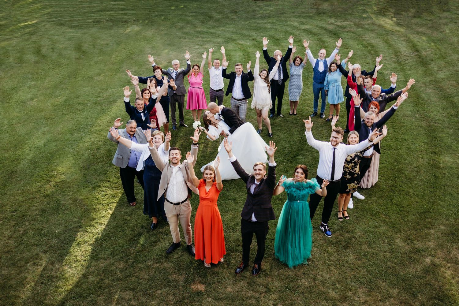 A large group of wedding guests form a heart shape on a green lawn while raising their arms in celebration.