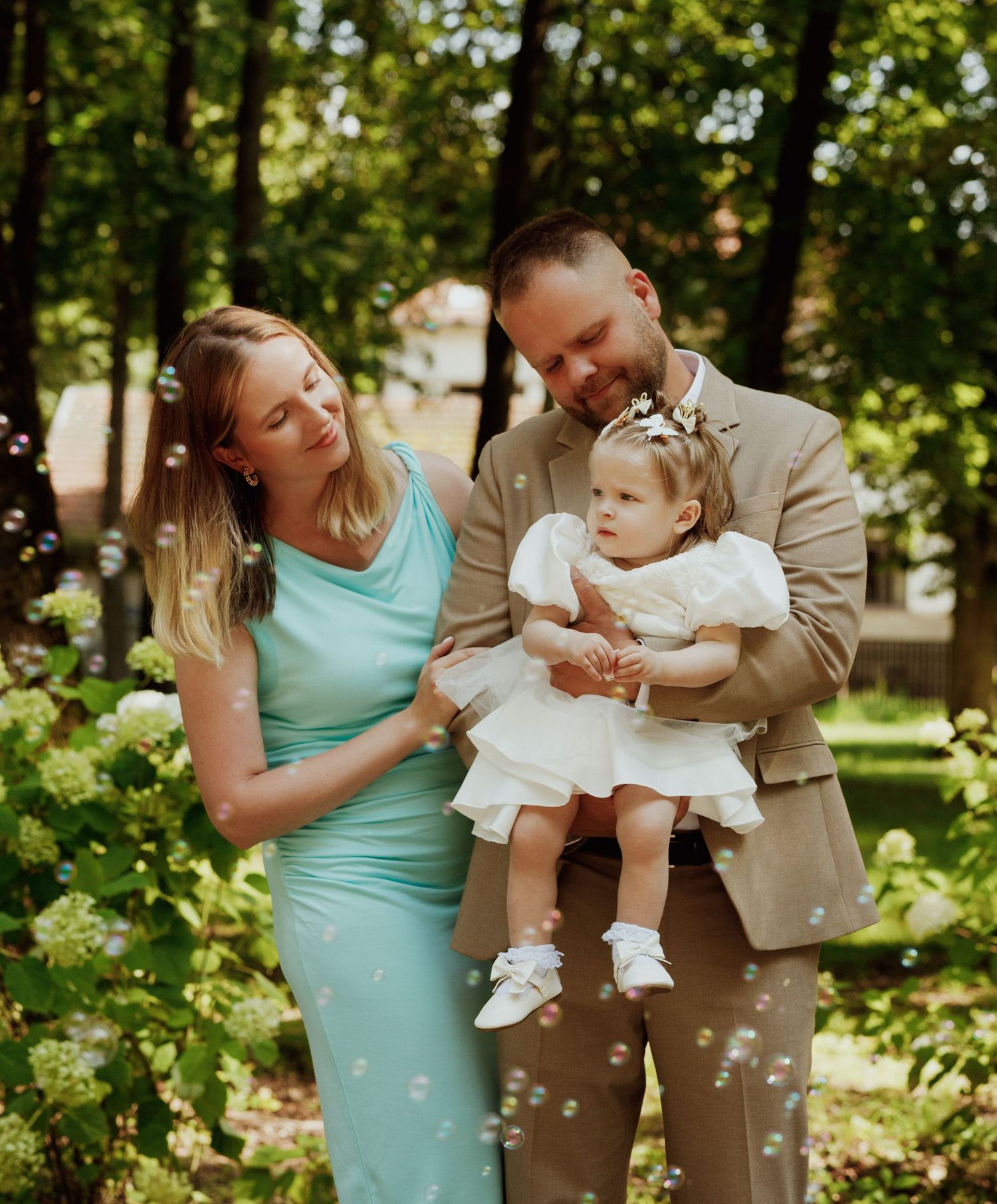 A couple in turquoise and tan outfits playfully interacts with their baby during an outdoor photo session in a sunlit garden.