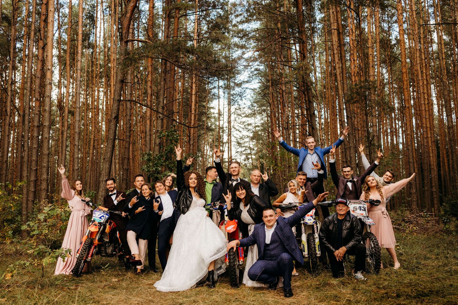 Wedding party group photo session in a scenic pine forest with guests posing together among tall trees.