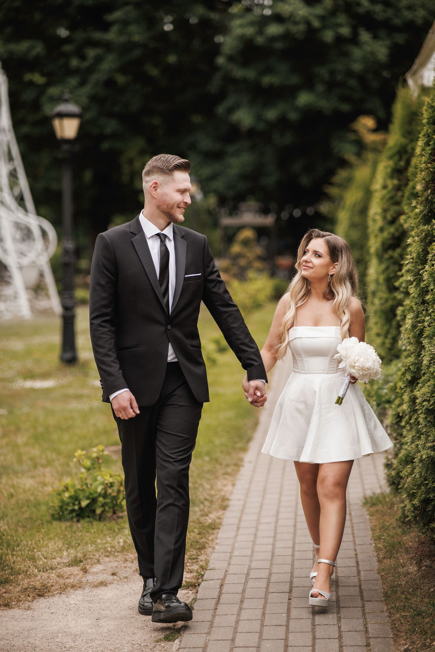 Couple in formal attire walking together on a garden path during a wedding photoshoot.