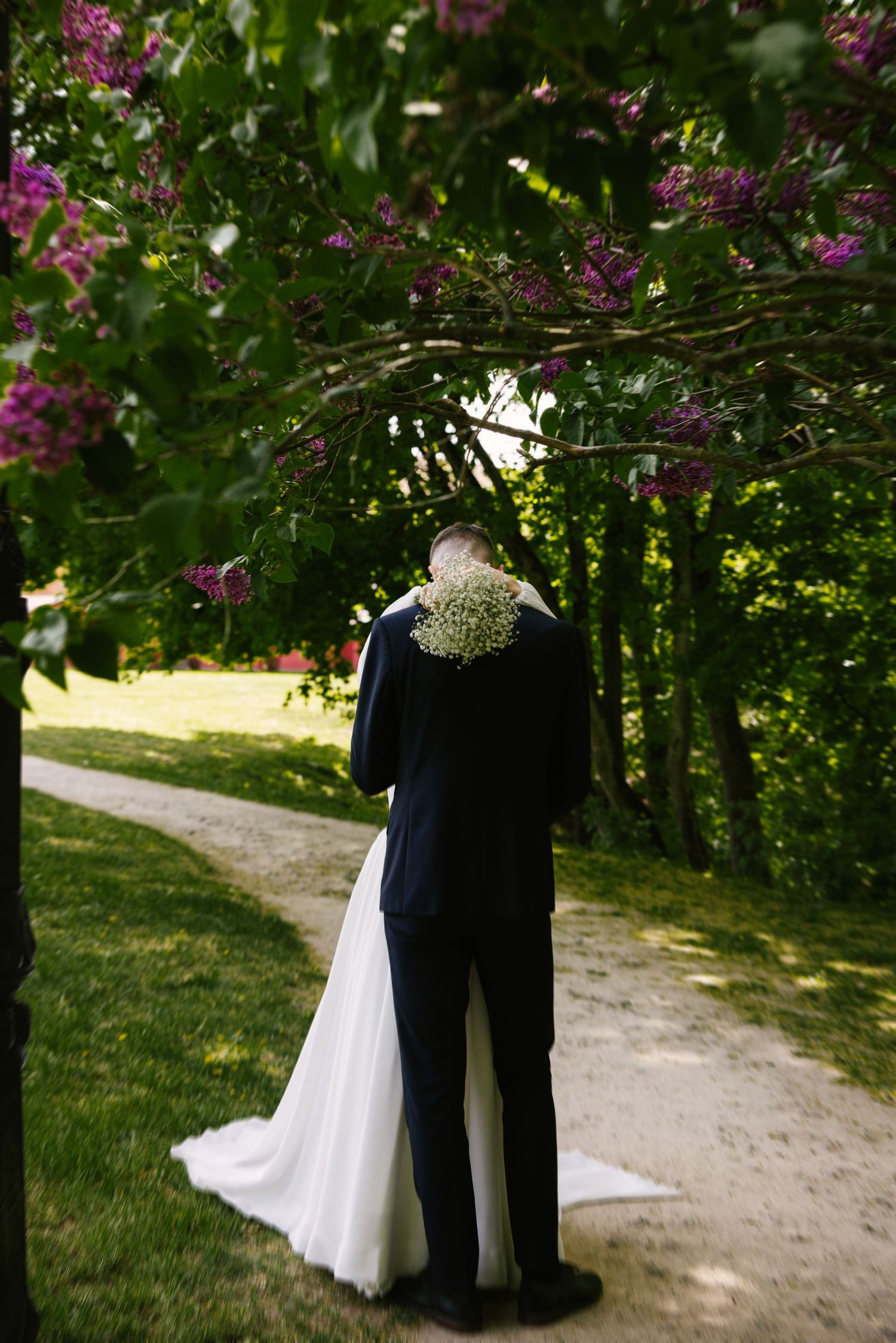 A wedding couple stands beneath flowering lilac branches on a garden path.