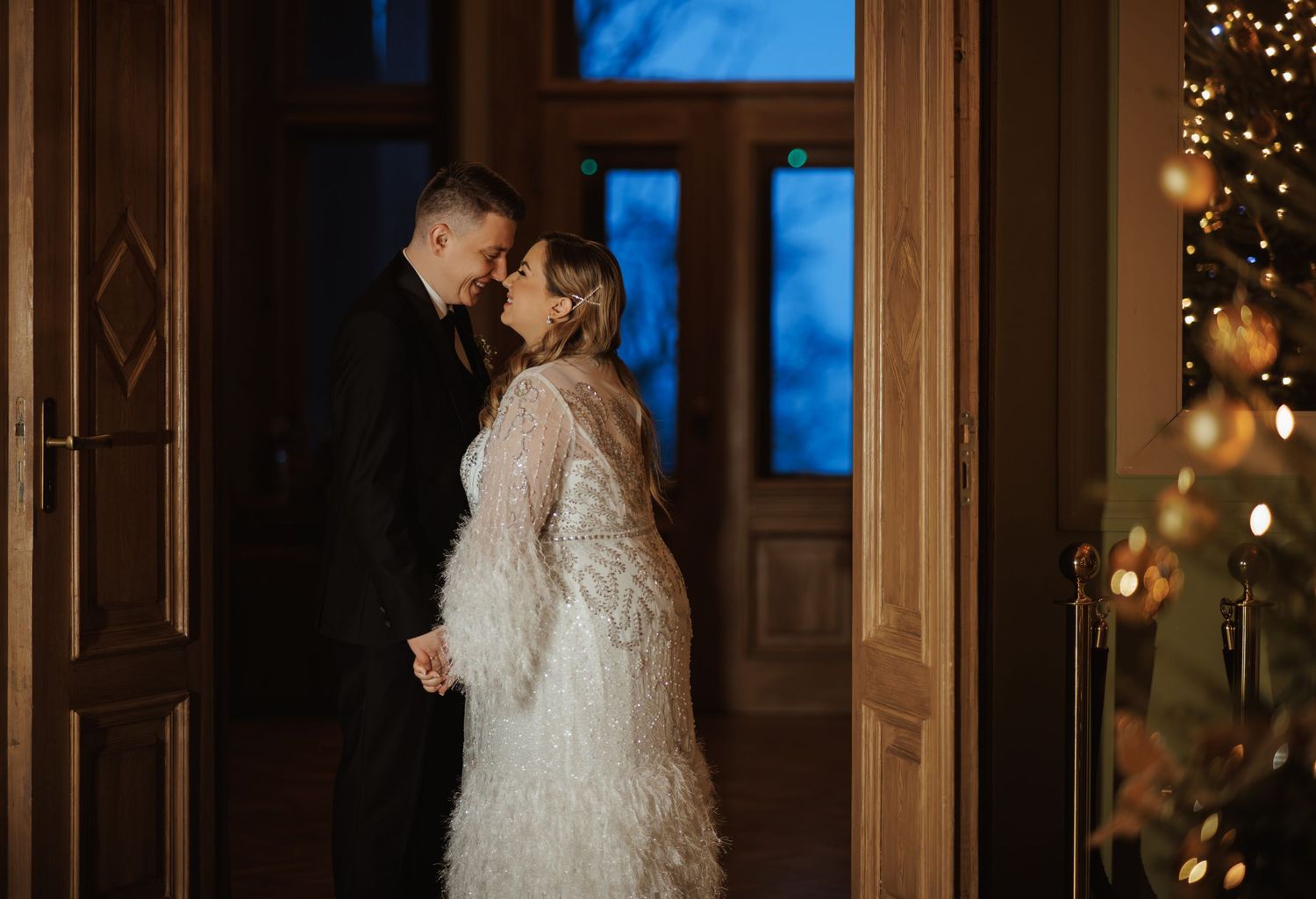 Elegant winter wedding scene with Christmas trees and twinkling lights in an ornate ballroom.